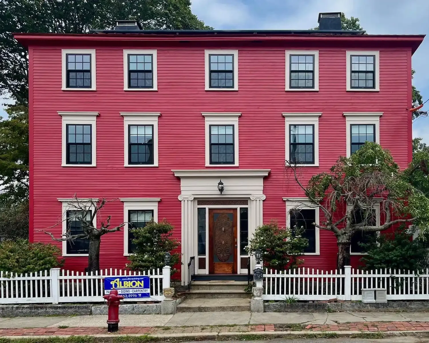 A three-story red house with white trim, black windows, and a wooden front door, surrounded by trees and a white picket fence.