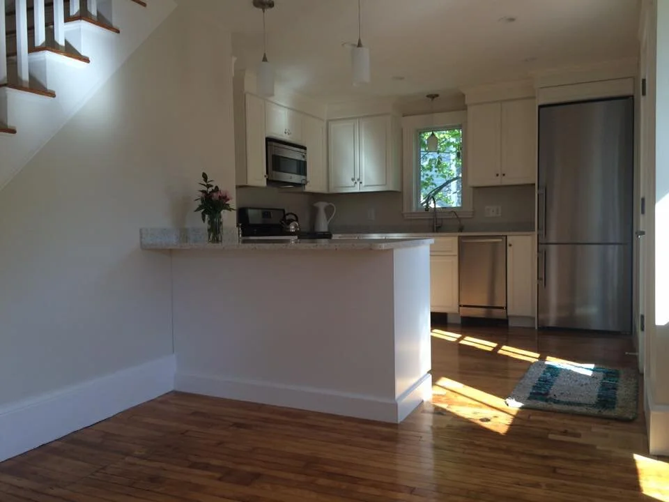 A bright kitchen with white cabinets in historic Newburyport. stainless steel refrigerator, and a small window, featuring hardwood floors and a kitchen island 