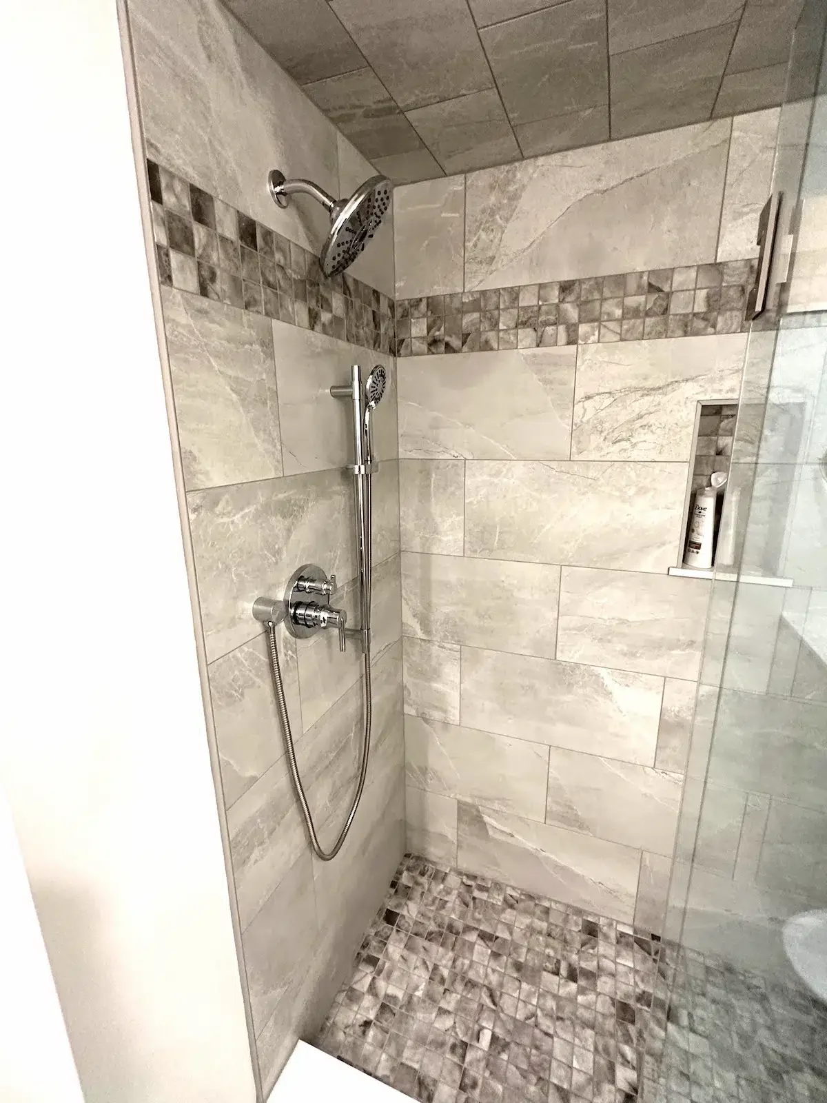 A modern walk-in shower with beige marble tiles on the walls, a strip of small square mosaic tiles, a built-in niche with a bottle of lotion, a rainfall showerhead, and a handheld showerhead with a hose.