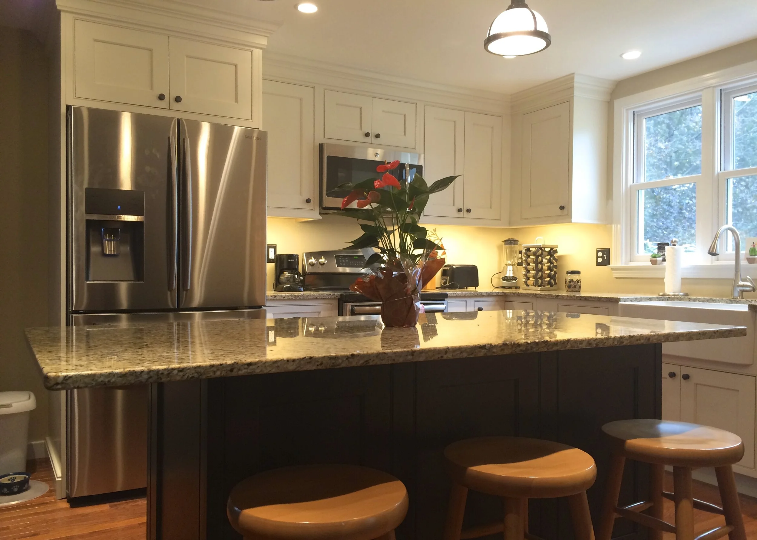 West Newbury Kitchen with stainless steel refrigerator, microwave, and granite countertop island with three wooden barstools, a flower arrangement, and a window overlooking trees.