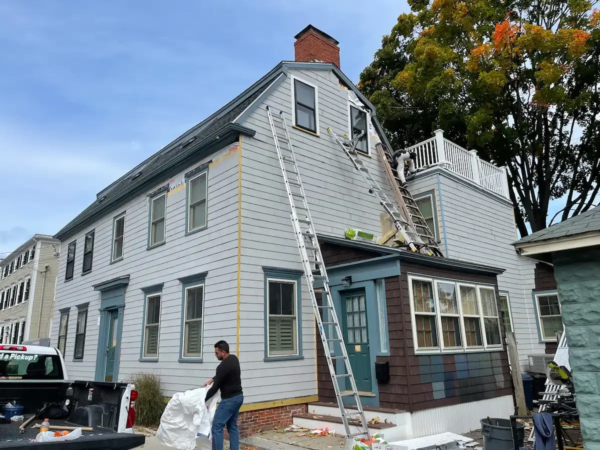 Workers renovate a multi-story house, removing or replacing siding with ladders positioned at various points on the house. A person is holding a large white bag, and a pickup truck is parked nearby.