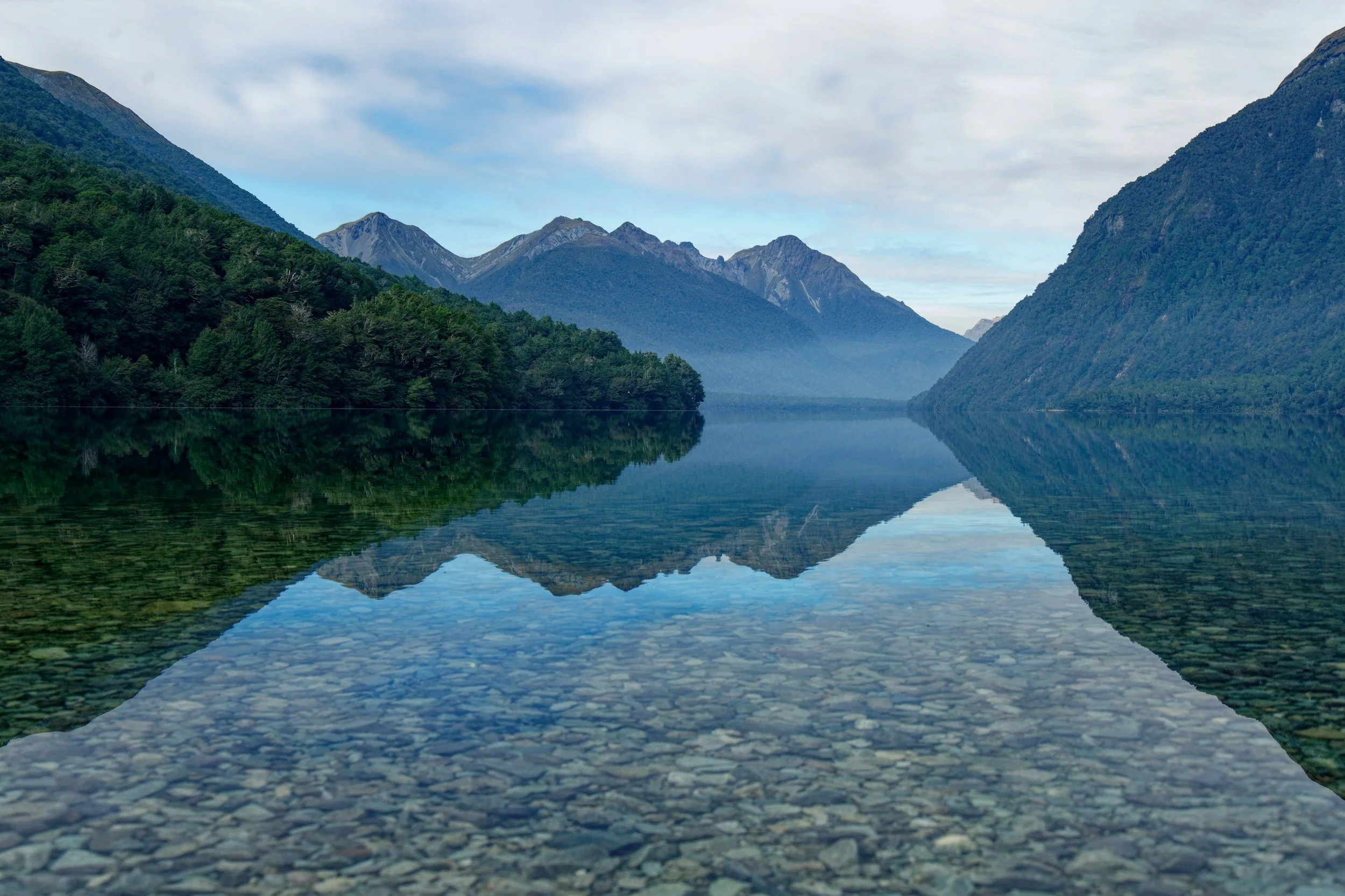Calm lake reflecting surrounding green mountains and distant peaks under a cloudy sky.