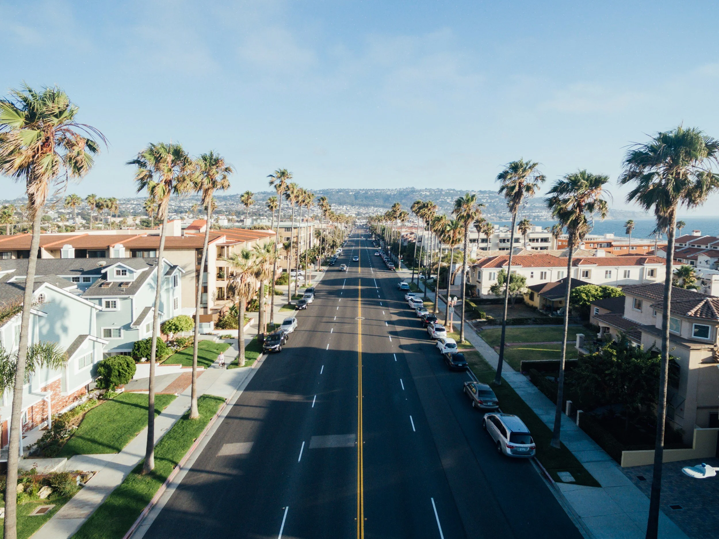 A sunny street lined with palm trees and residential houses leading to the ocean in the distance.