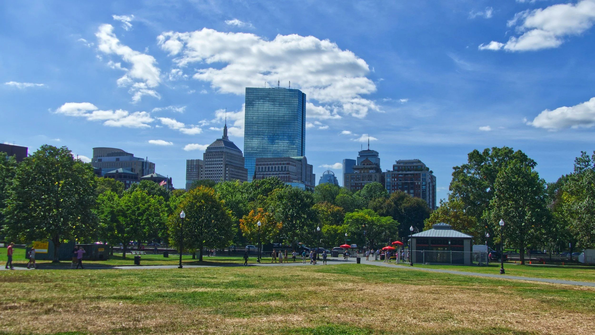 City skyline with tall modern buildings, green trees, and a park with people walking under a partly cloudy blue sky.