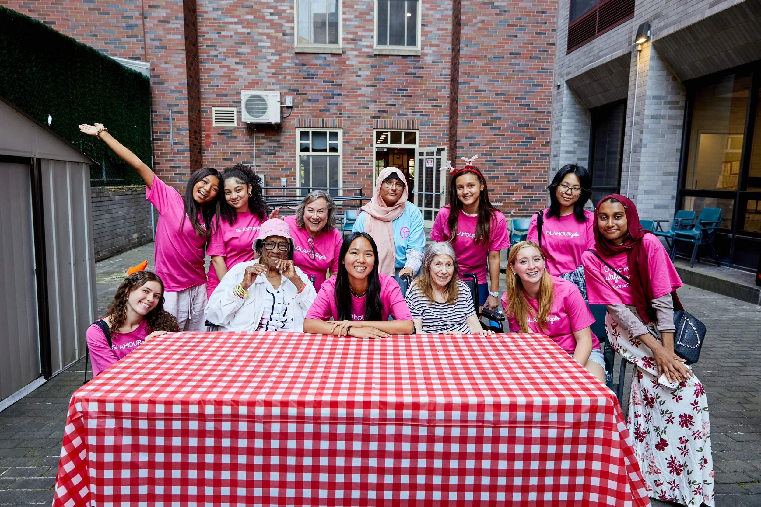 Group of diverse women and girls, some with disabilities, gathered around a table with a red and white checkered tablecloth outdoors, smiling for a photo at a social event.