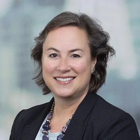 Professional woman with short dark hair, wearing a dark blazer and necklace, smiling against a blurred light-colored background.