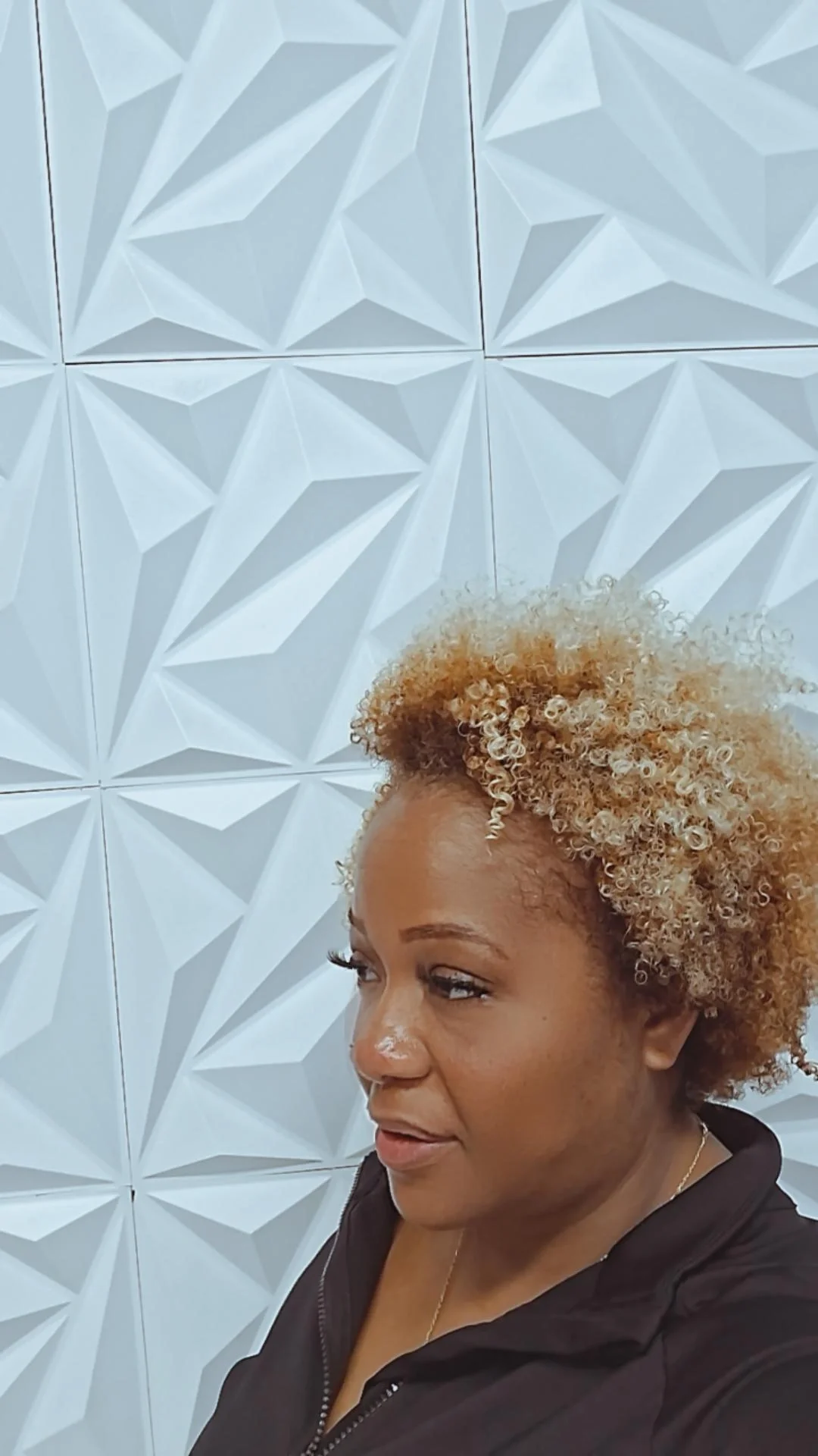 A woman with curly blonde hair takes a selfie in front of a white textured wall with geometric patterns.
