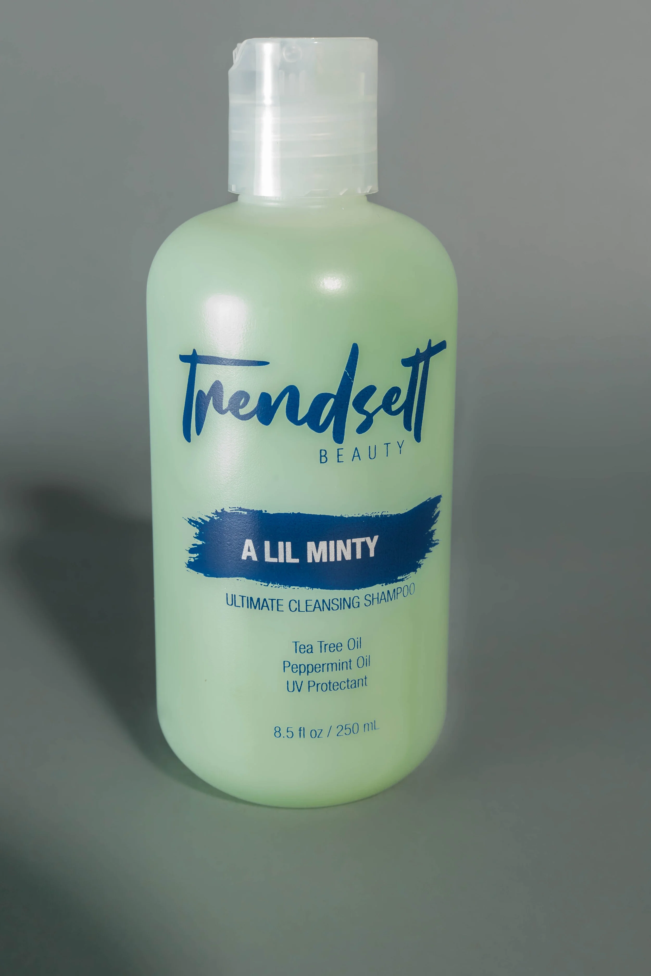 A bottle of Trendsett Beauty A Lil Minty ultimate cleansing shampoo, with tea tree oil, peppermint oil, and UV protectant, in a light green bottle with blue text on a gray background.