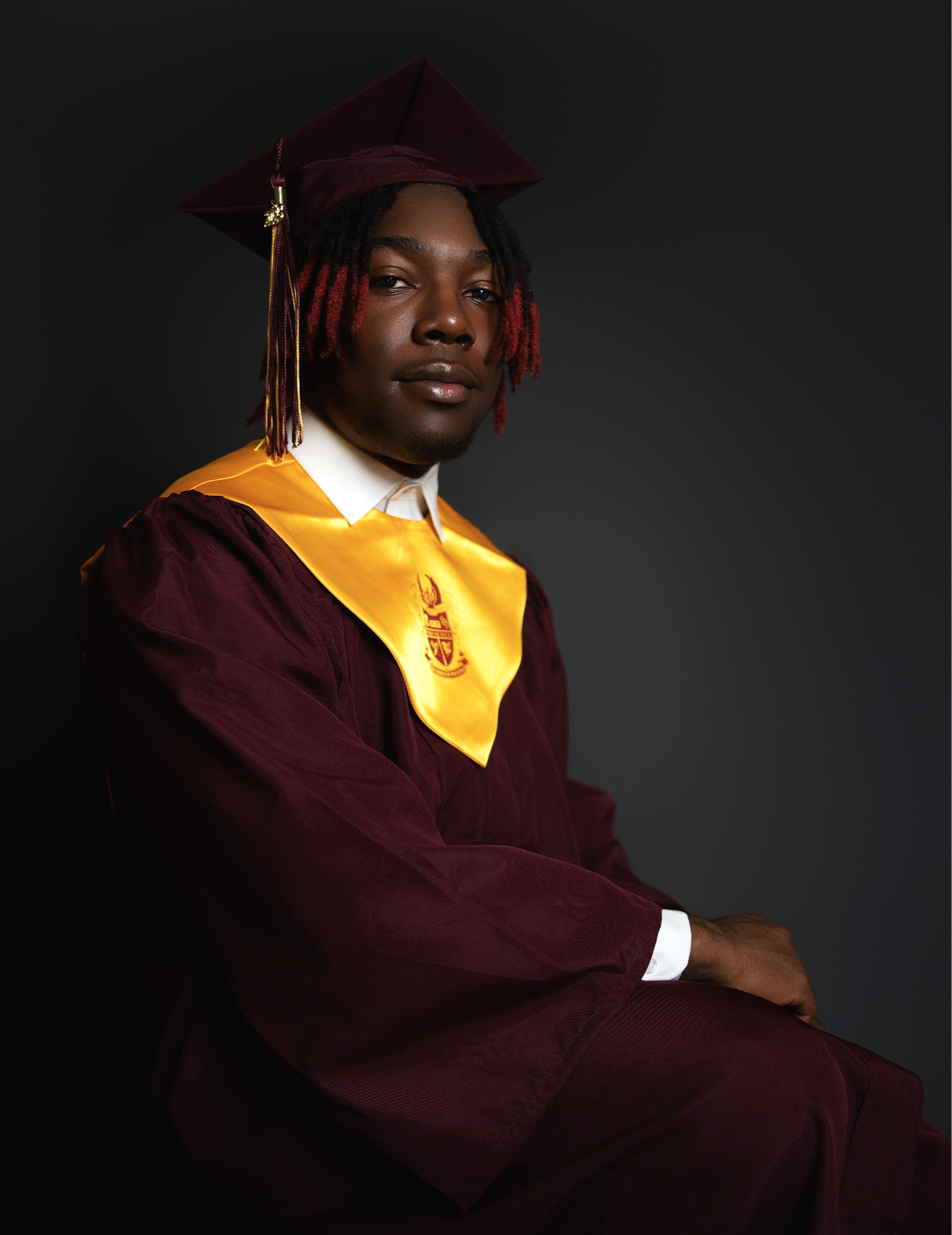A young person in graduation attire, wearing a maroon cap and gown with a yellow stole, sitting against a dark background.