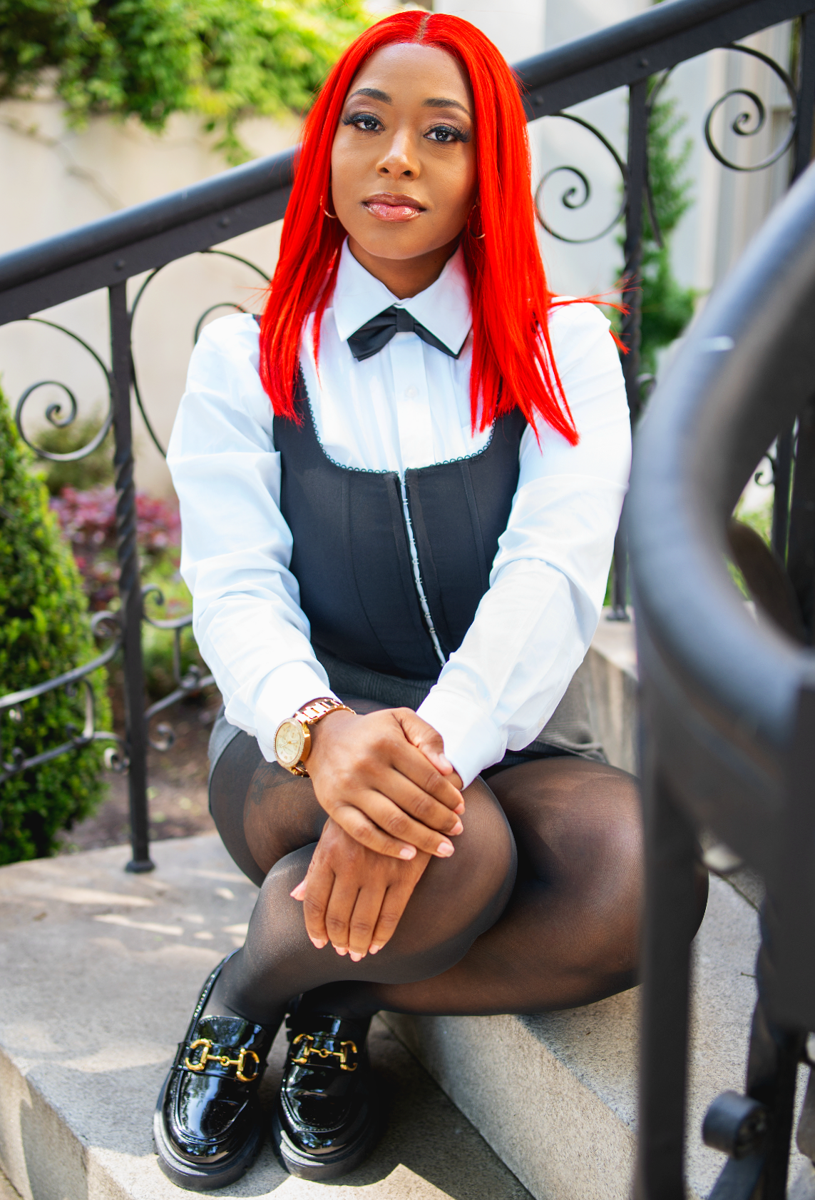 Woman with bright red hair sitting on outdoor stairs, wearing a white shirt, black vest with zipper, black sheer tights, black loafers with gold hardware, and gold watch, posing with her hands clasped on her knee.