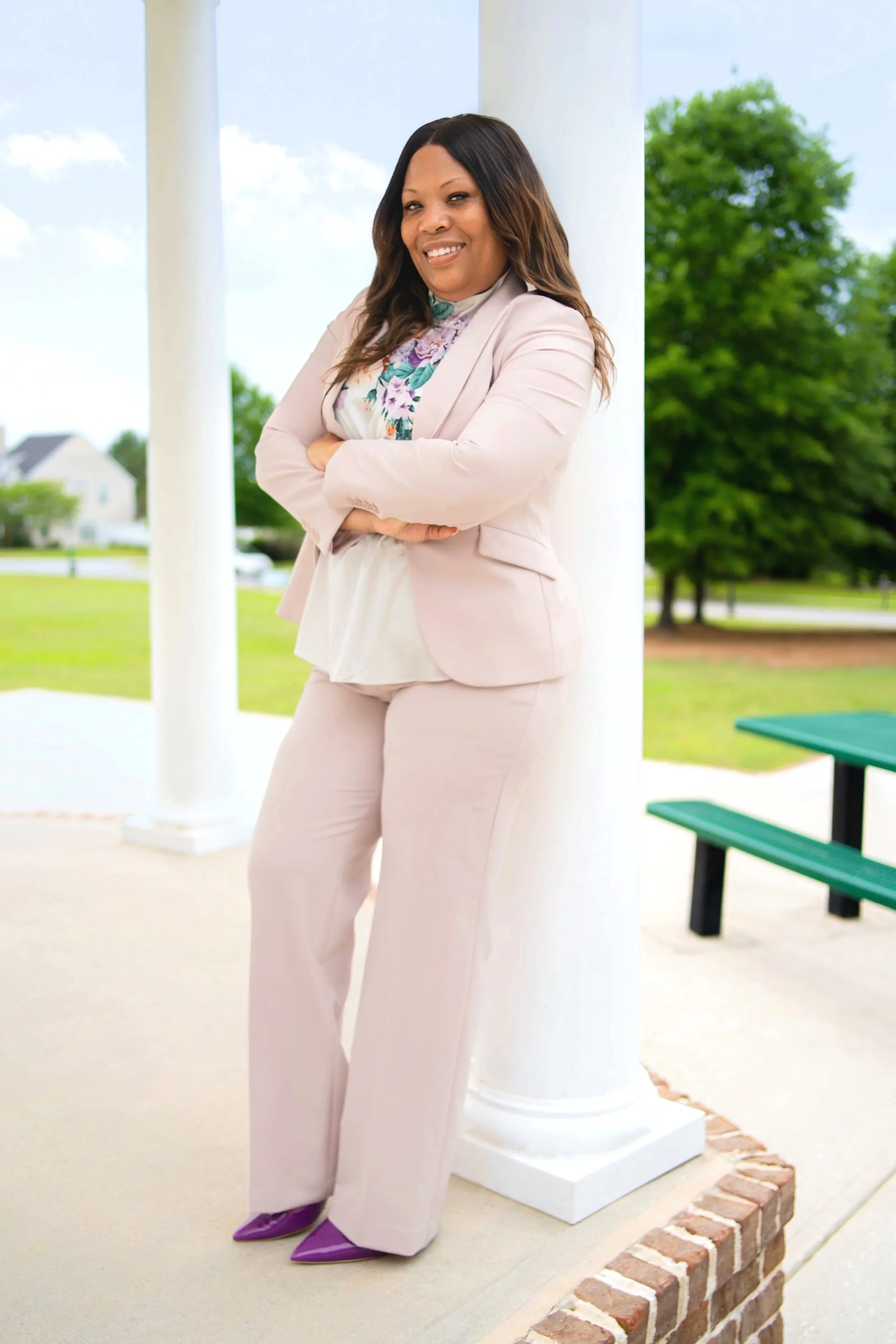 A woman standing outdoors, leaning against a white pillar, wearing a light pink suit, floral blouse, and purple shoes, smiling with arms crossed on a bright day with trees and a house in the background.