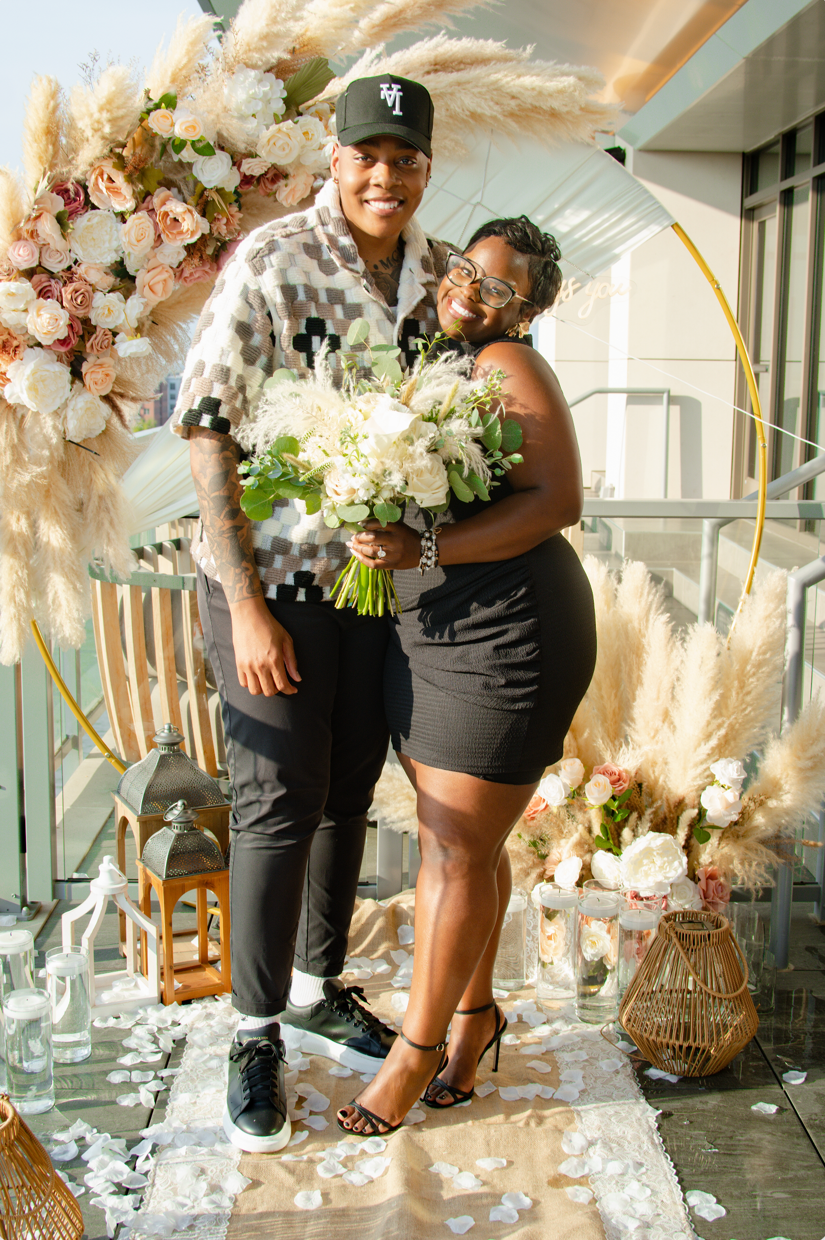 A person in a baseball cap and plaid shirt stands beside a person in a black dress, holding a bouquet of white flowers, at a daytime outdoor proposal celebration decorated with flowers, pampas grass, candles, and lanterns.