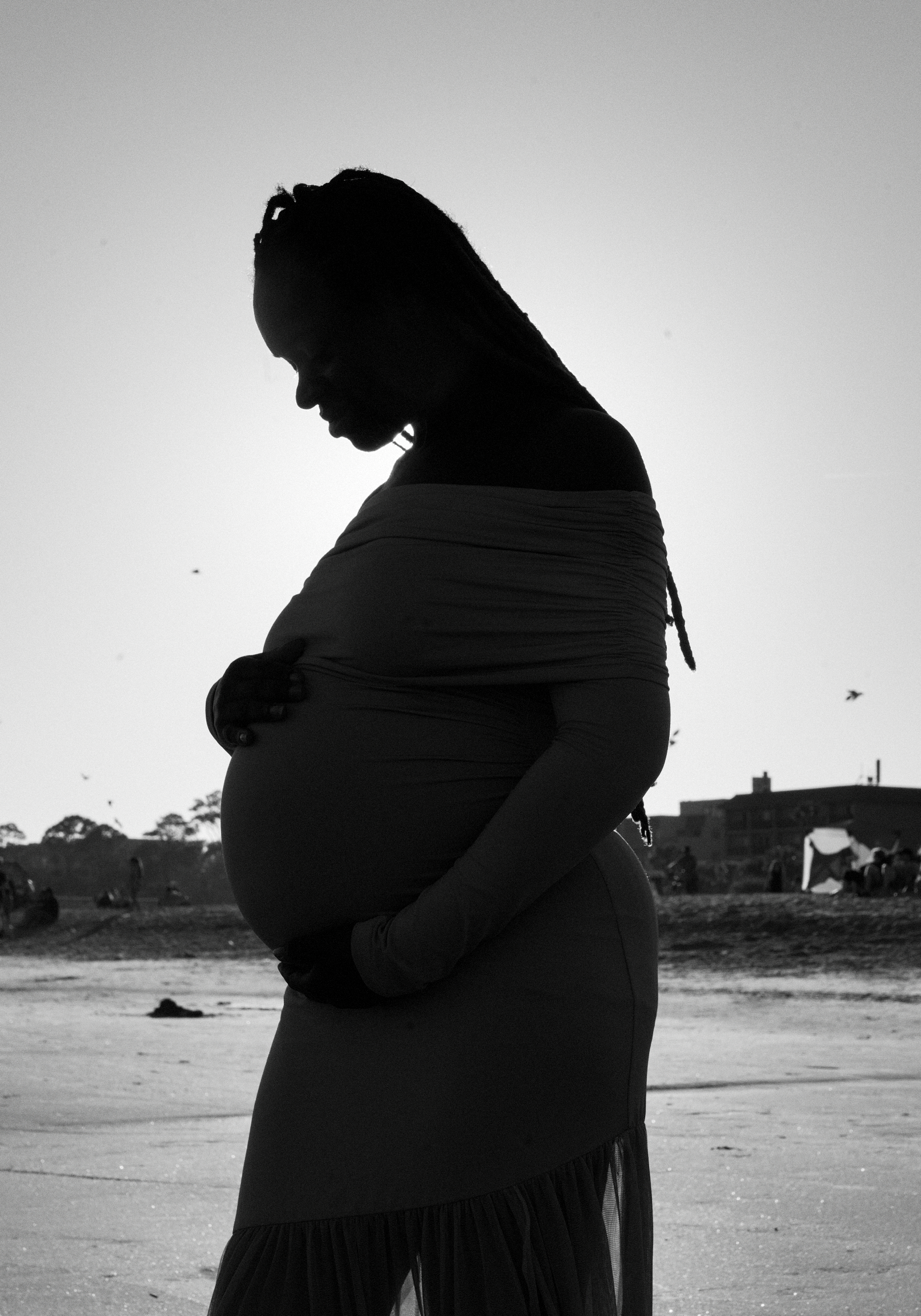Silhouette of a pregnant woman standing on a beach with her arms crossed, looking down, with birds flying in the sky and buildings in the background.