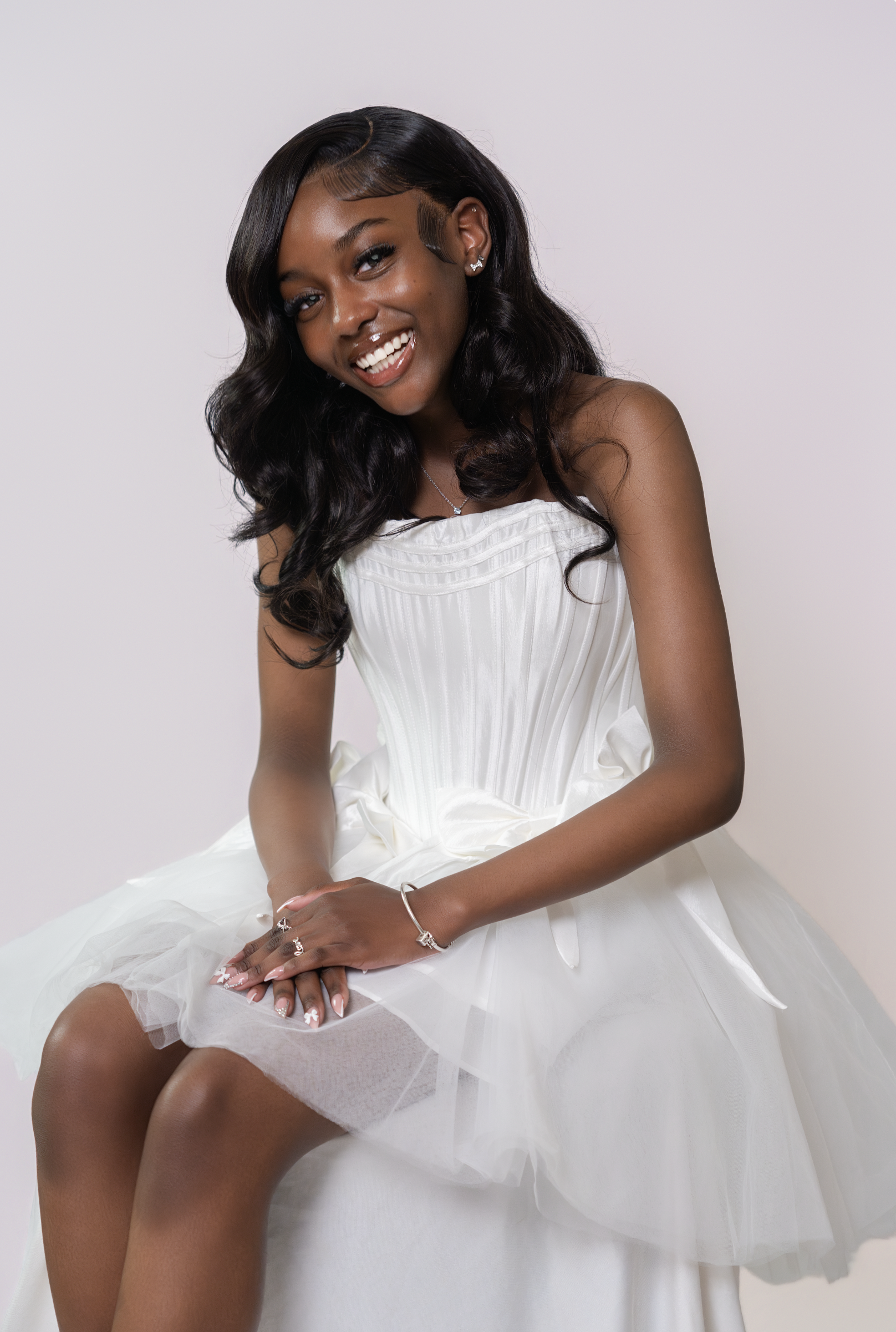 A young woman wearing a white dress with black wavy hair, smiling, sitting against a light background.