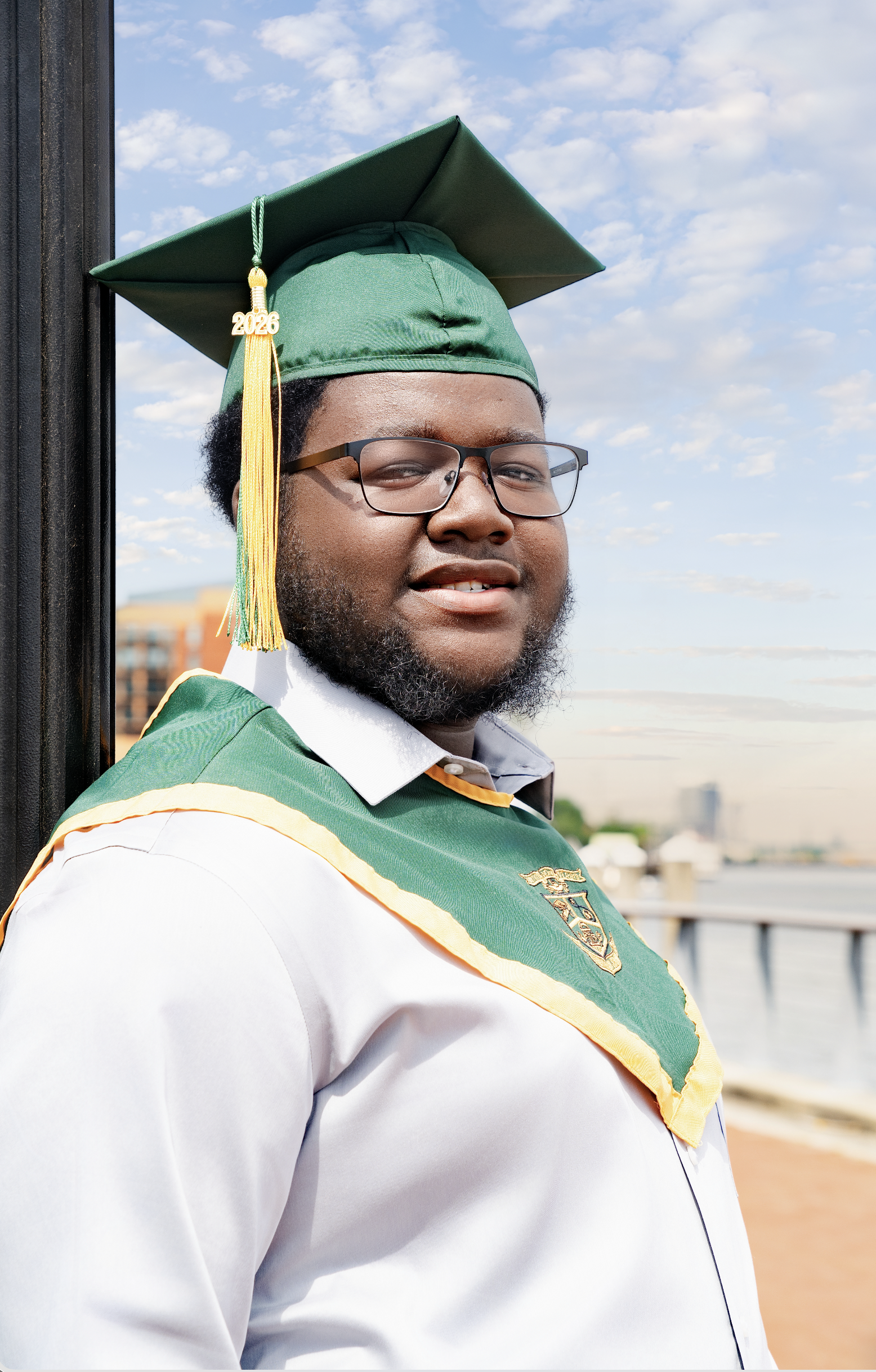 Graduate in green cap and gown with a yellow tassel marked 2026, wearing glasses, standing outdoors near a body of water under a partly cloudy sky.