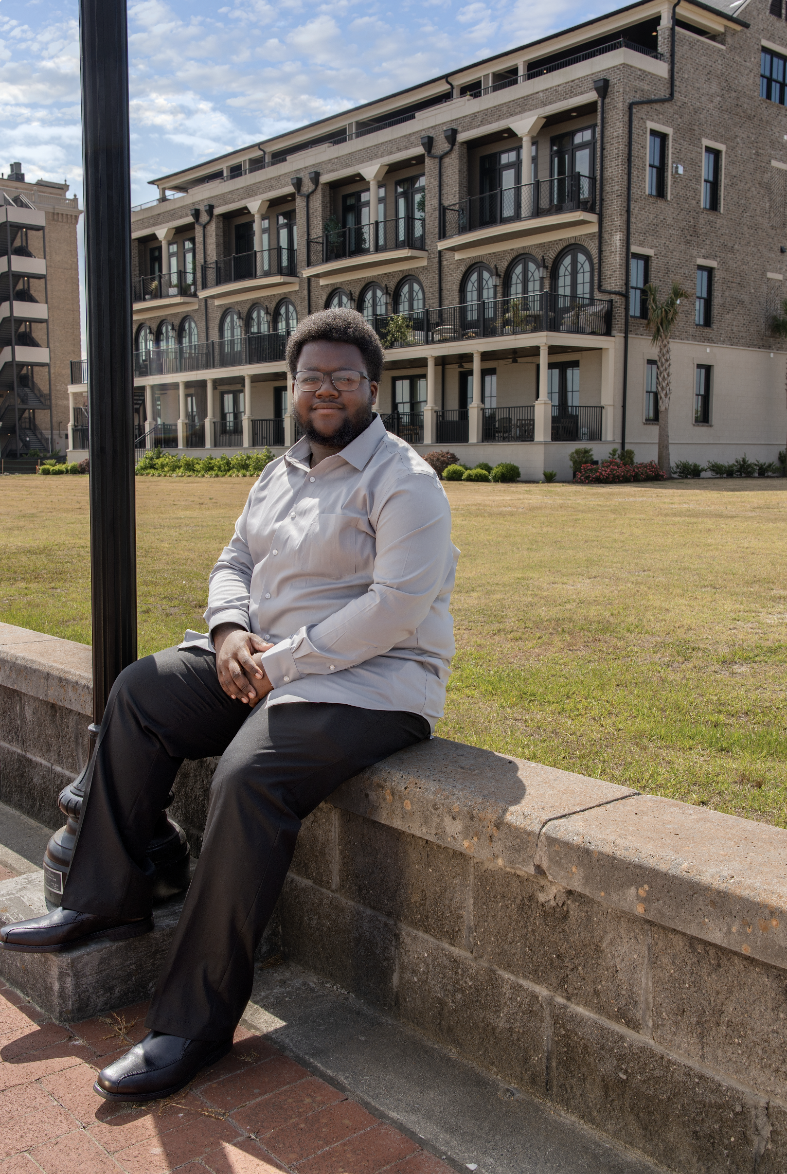 Young man with glasses and beard sitting on a stone ledge outdoors with an apartment building and palm tree in the background.