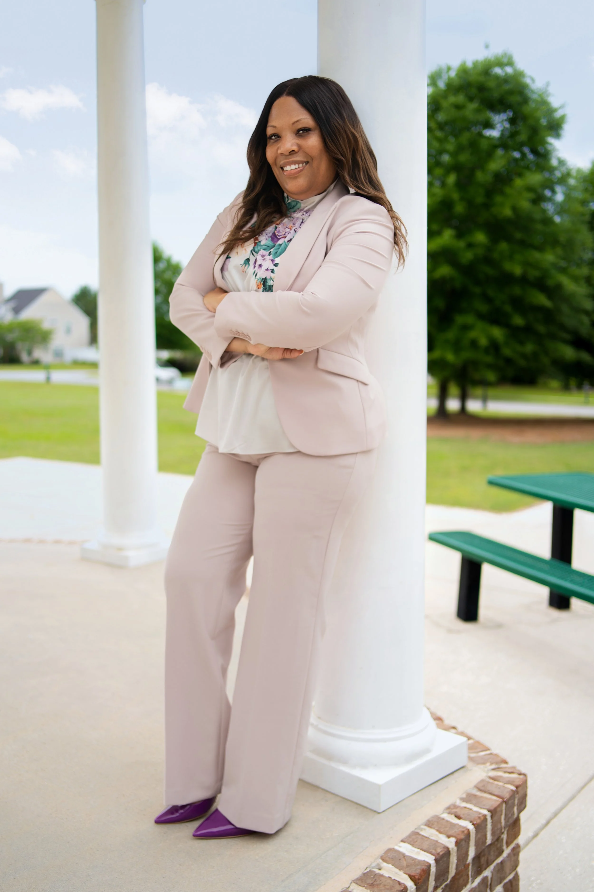 A woman standing outdoors, leaning against a white pillar, wearing a light pink suit, floral blouse, and purple shoes, smiling with arms crossed on a bright day with trees and a house in the background.