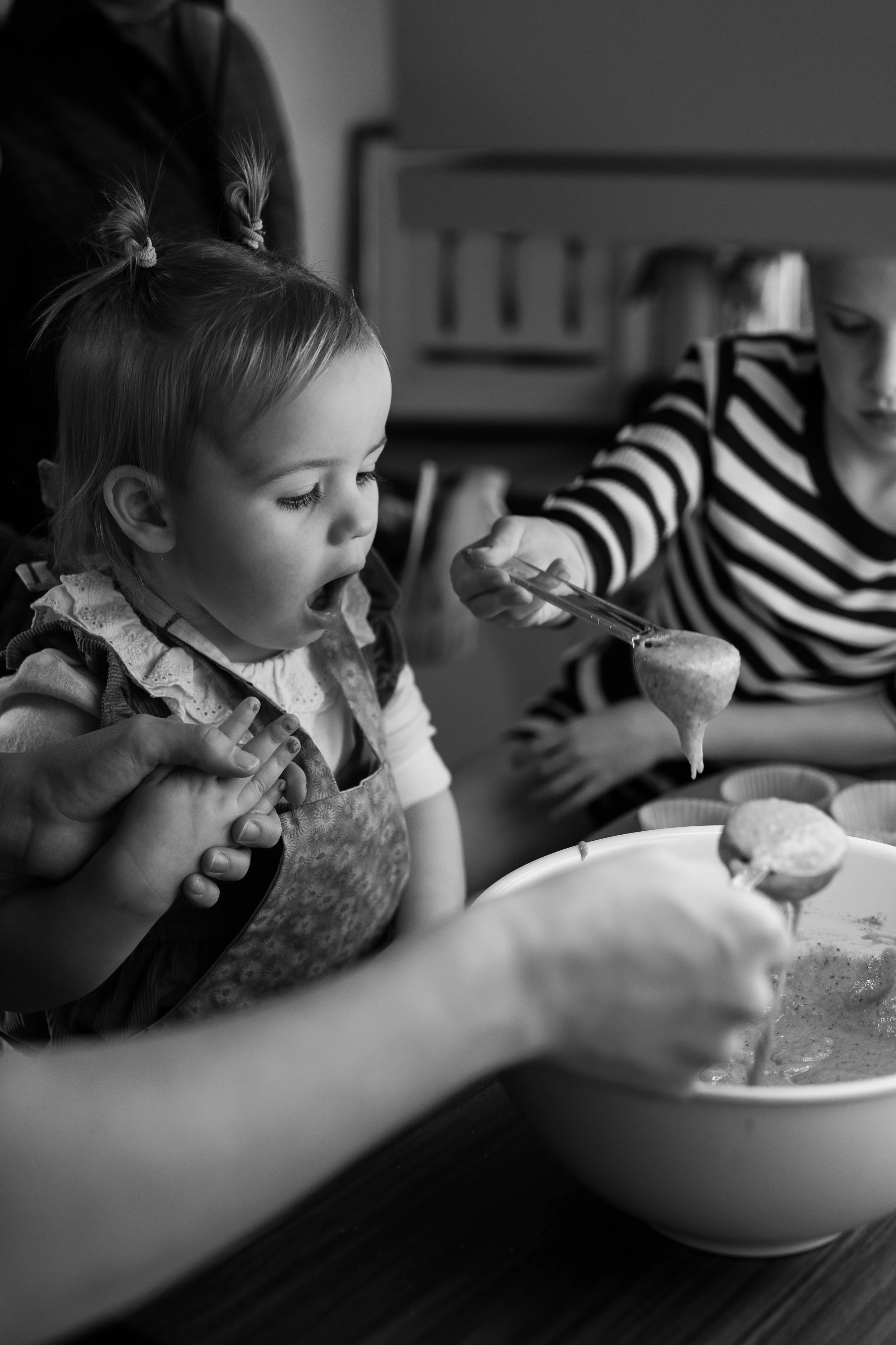 A young girl eagerly opens her mouth to eat a food ball from a spoon held by a woman in a striped shirt. Another person is guiding her hands, while a bowl of food is in front of them.