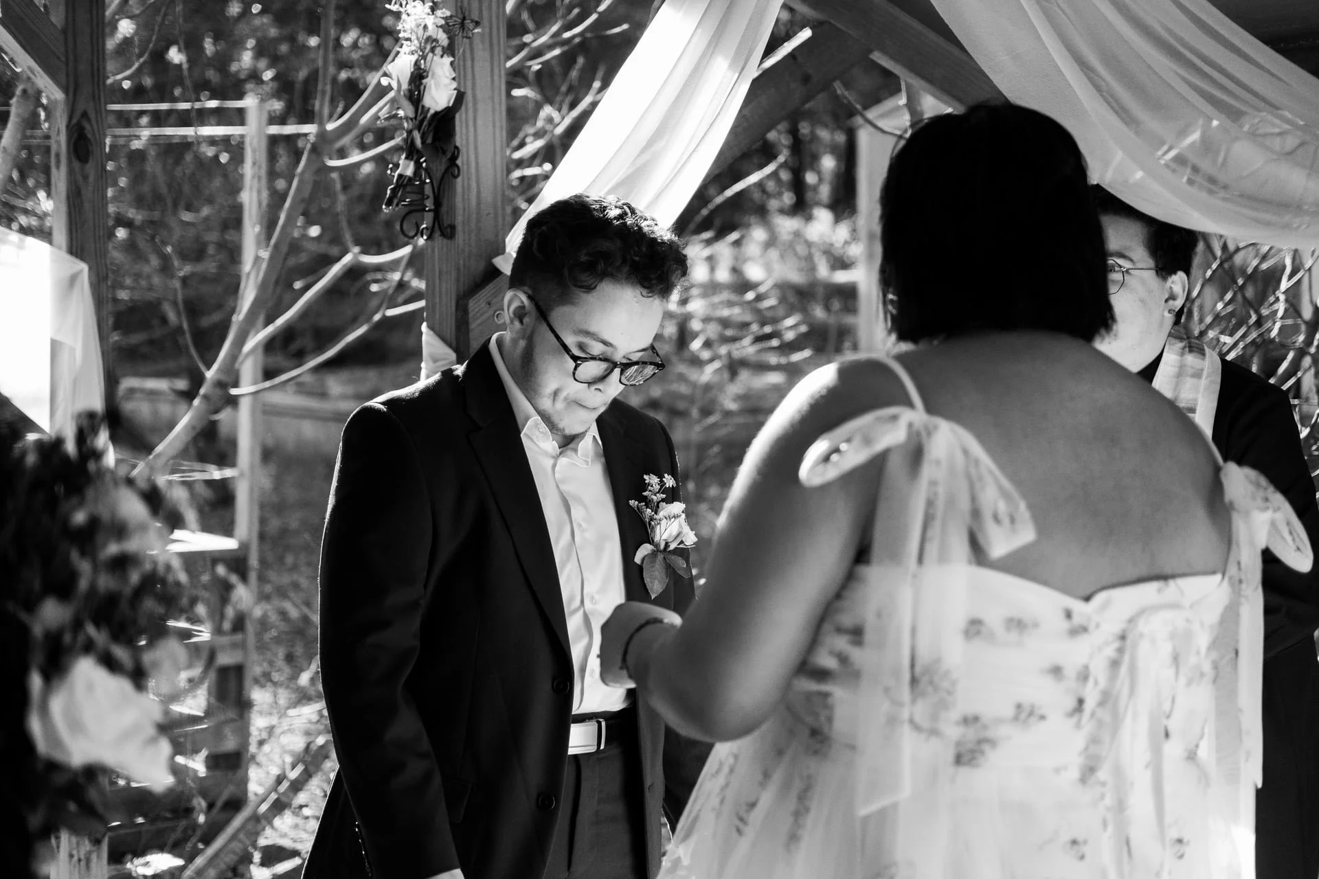 A black and white photo of a wedding ceremony outdoors with three people participating, one man in a suit with glasses and a woman in a dress with a floral pattern, and another person with short dark hair and glasses.