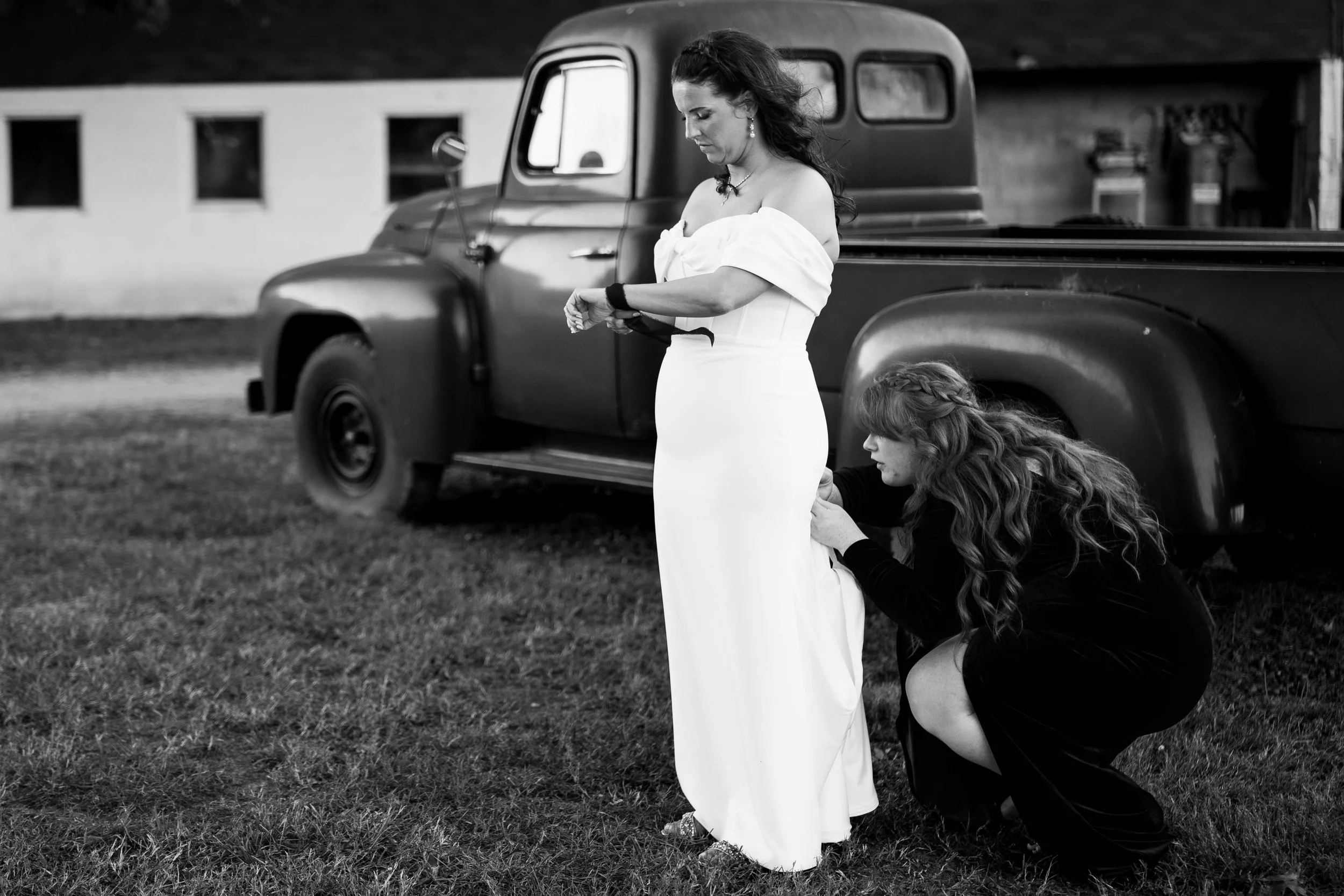A woman in an elegant white dress is being assisted by another woman in black as she prepares for a formal event, standing beside a vintage truck outdoors.