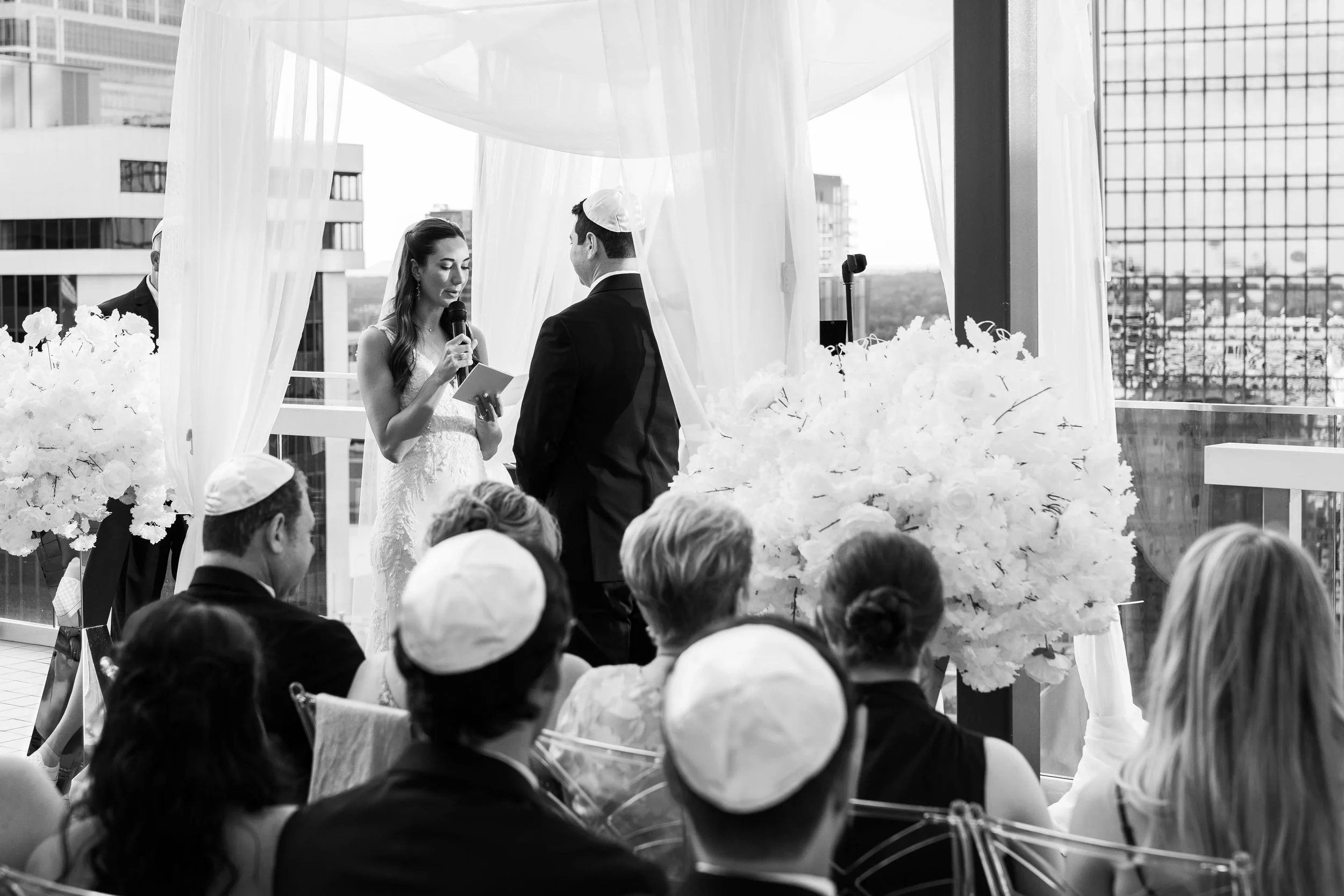 A couple having a wedding ceremony on a rooftop, with the bride reading vows and the groom listening, surrounded by guests wearing kippahs and seated in front of an urban cityscape.