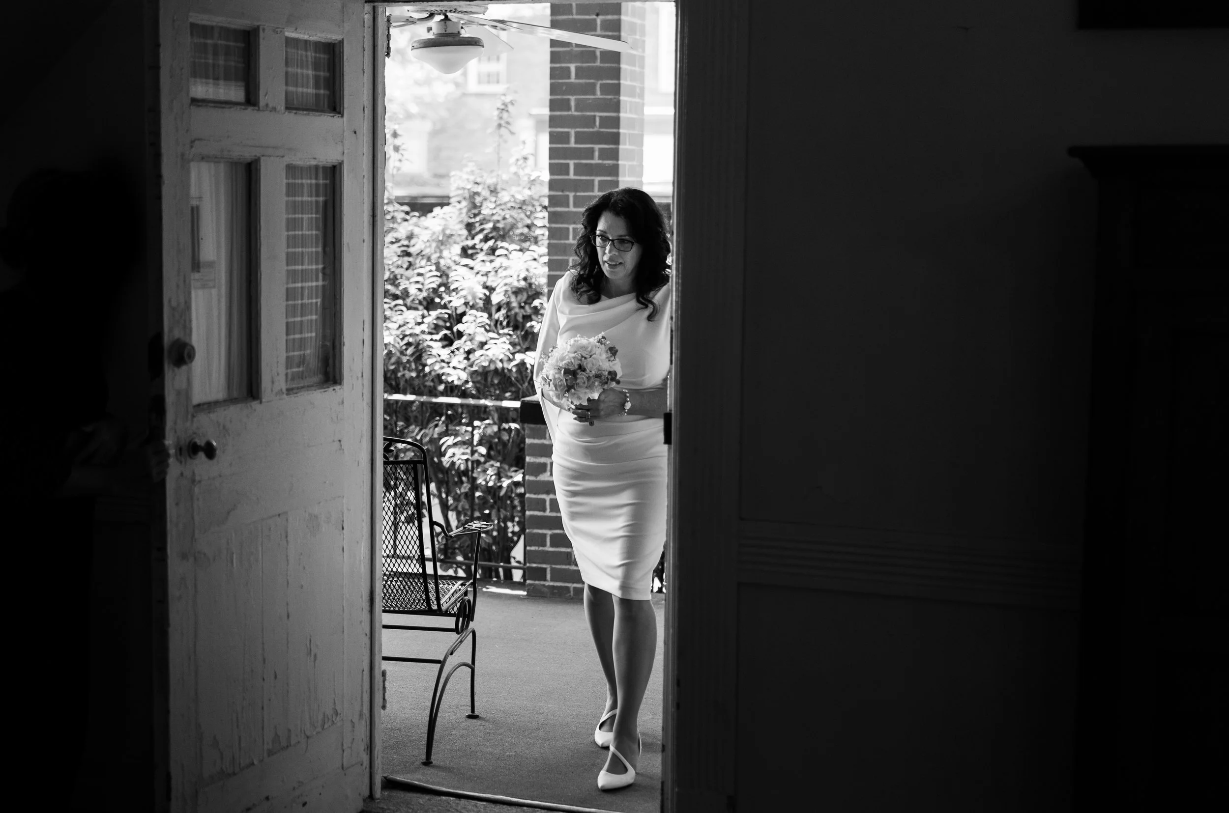 A woman in a white dress holding a bouquet, standing on a balcony outside a building, with an outdoor background of plants and brick. The photo is black and white.