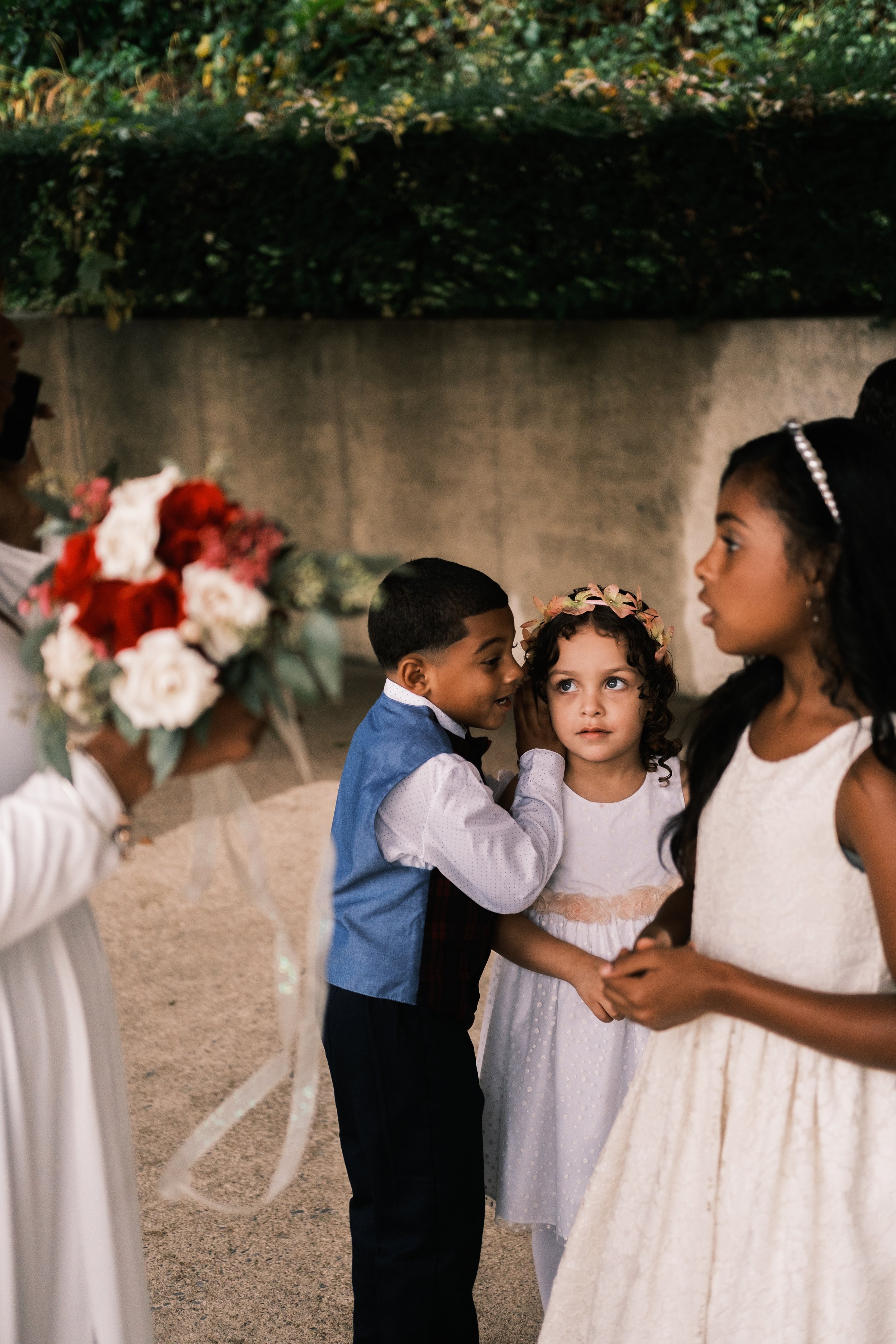 Young boy dressed in a tux and a flower girl at a wedding. the young boy is whispering into the ear of the flower girl during a wedding in Charlotte.