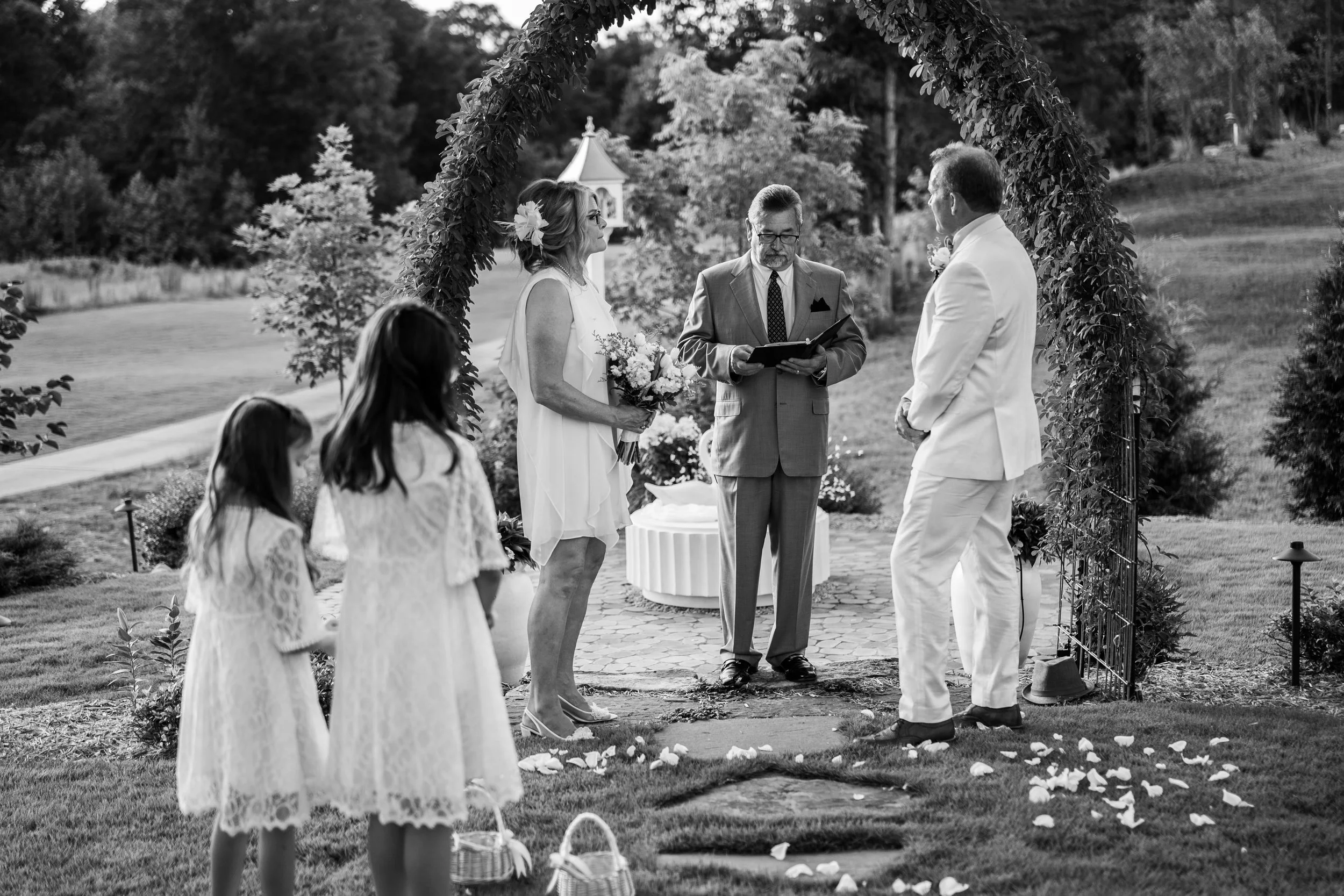 A black-and-white photo of a wedding ceremony outdoors with a couple standing before an officiant under an arch decorated with greenery and flowers. Two flower girls in lace dresses are in the foreground, holding baskets. The ceremony takes place in 