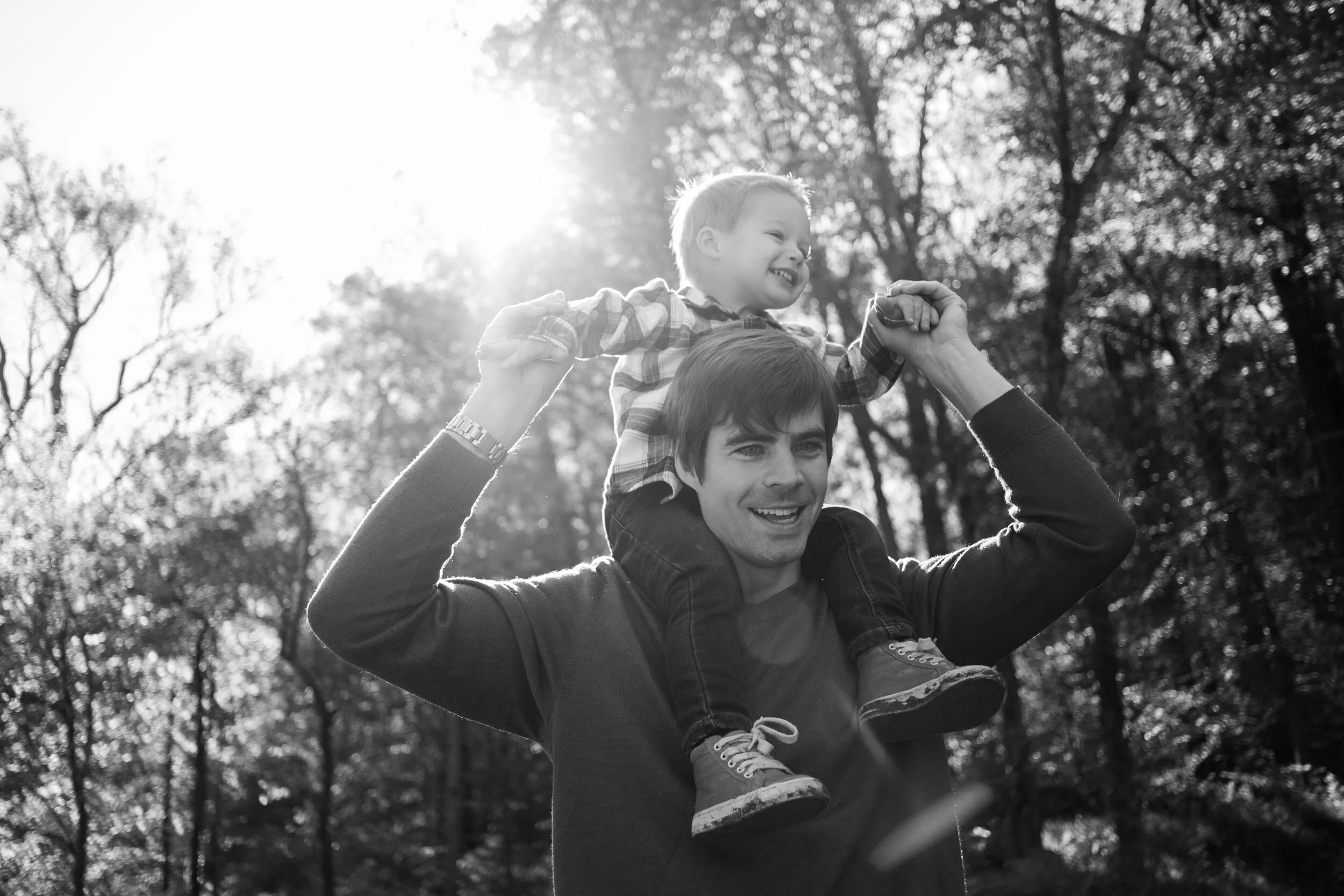 A man carrying a young boy on his shoulders outdoors in a park with trees, sunlight, and a bright sky in the background, captured in black and white.