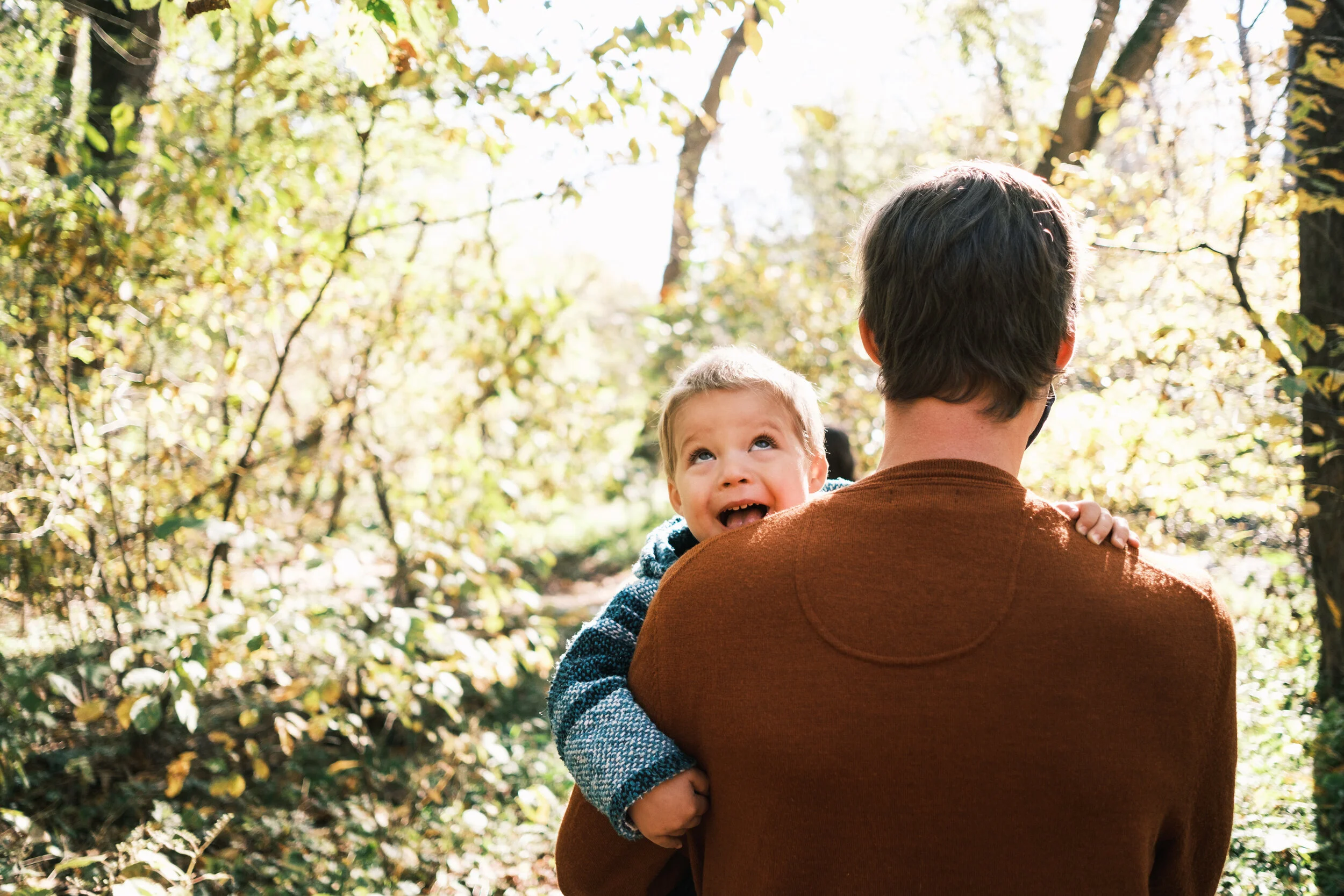 A father holding his young son in an outdoor setting with trees and sunlight filtering through the leaves during a family photo shoot in charlotte
