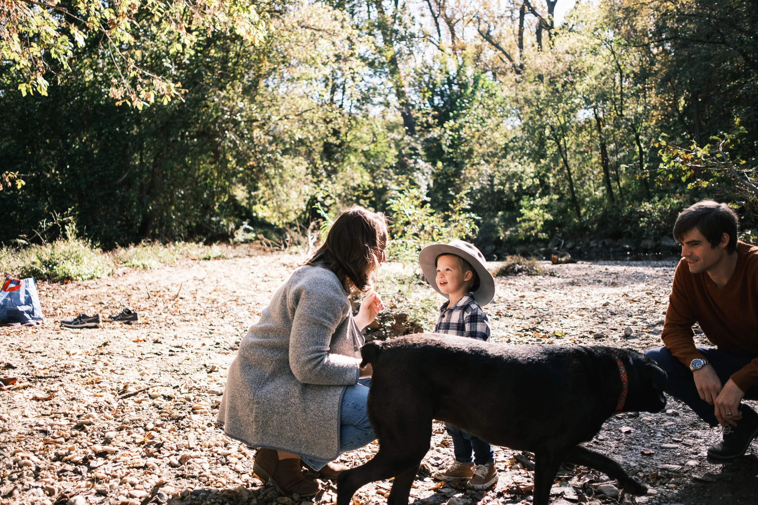 Family of three with a black dog enjoying a day outdoors in a wooded area, with sunlight filtering through trees at a family photo shoot at Lake Wylie.
