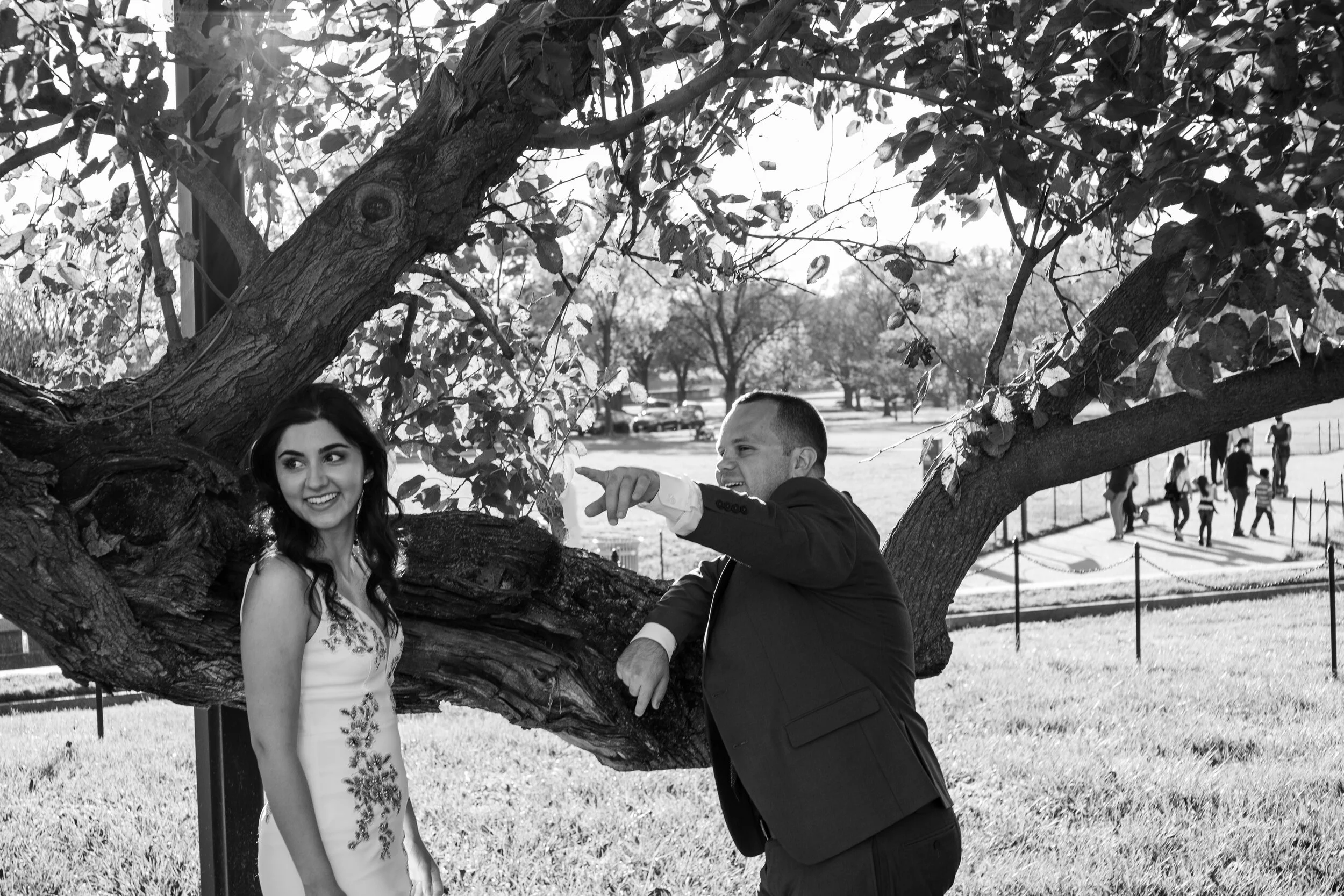 A smiling woman in a floral dress stands beside a man in a suit under a large tree. The man is pointing and talking to her. There are other people walking in the background in a park.