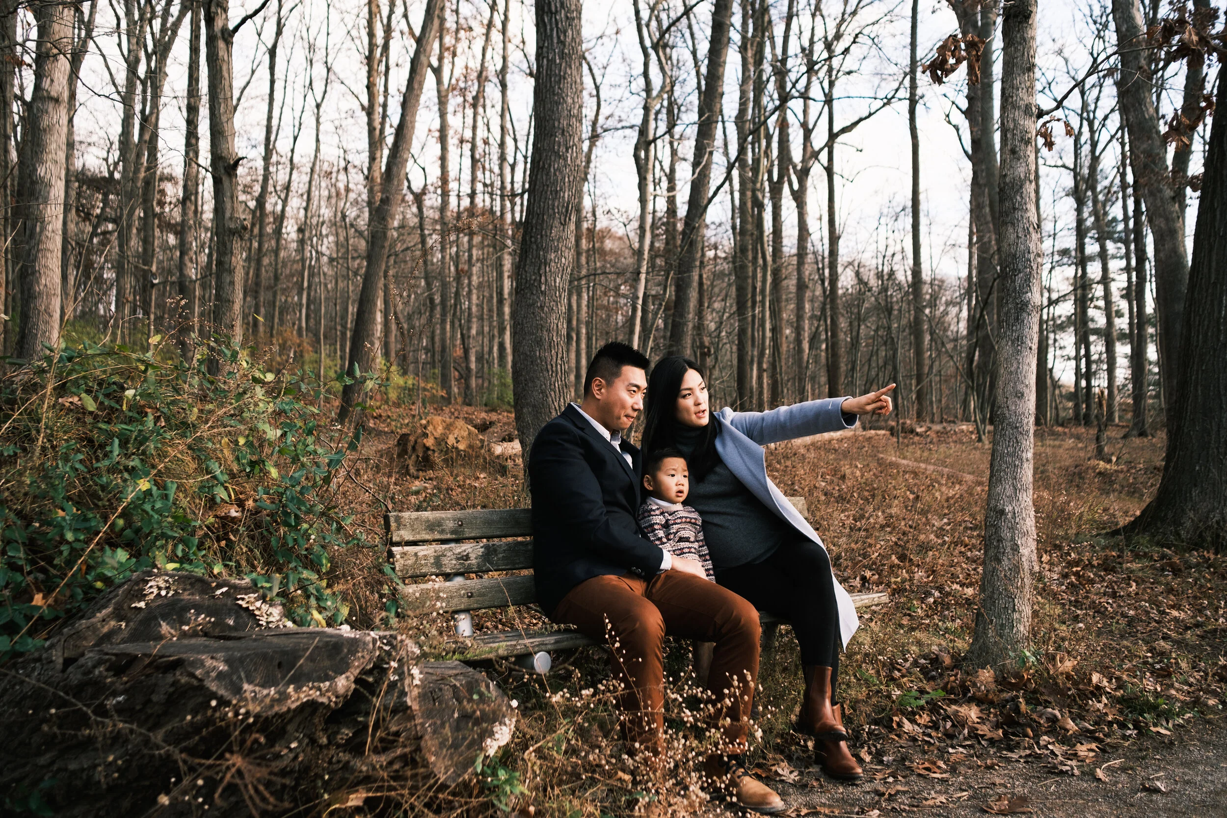 A family of three sitting on a park bench in a wooded area, with leafless trees and dry leaves on the ground, during late autumn or winter at Lake Wylie for a family photo session.