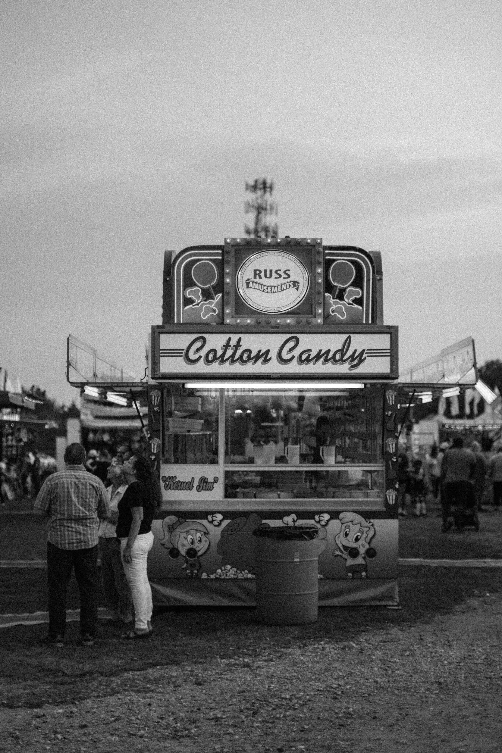 Black and white image of a cotton candy stand at a fair with people gathered around