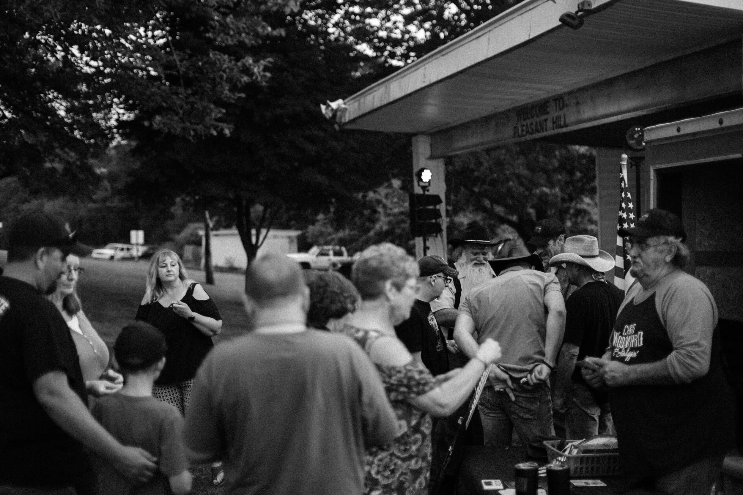 Group of people socializing outdoors in a park setting near a building with a banner reading 'Welcome to Pleasant Hill.'