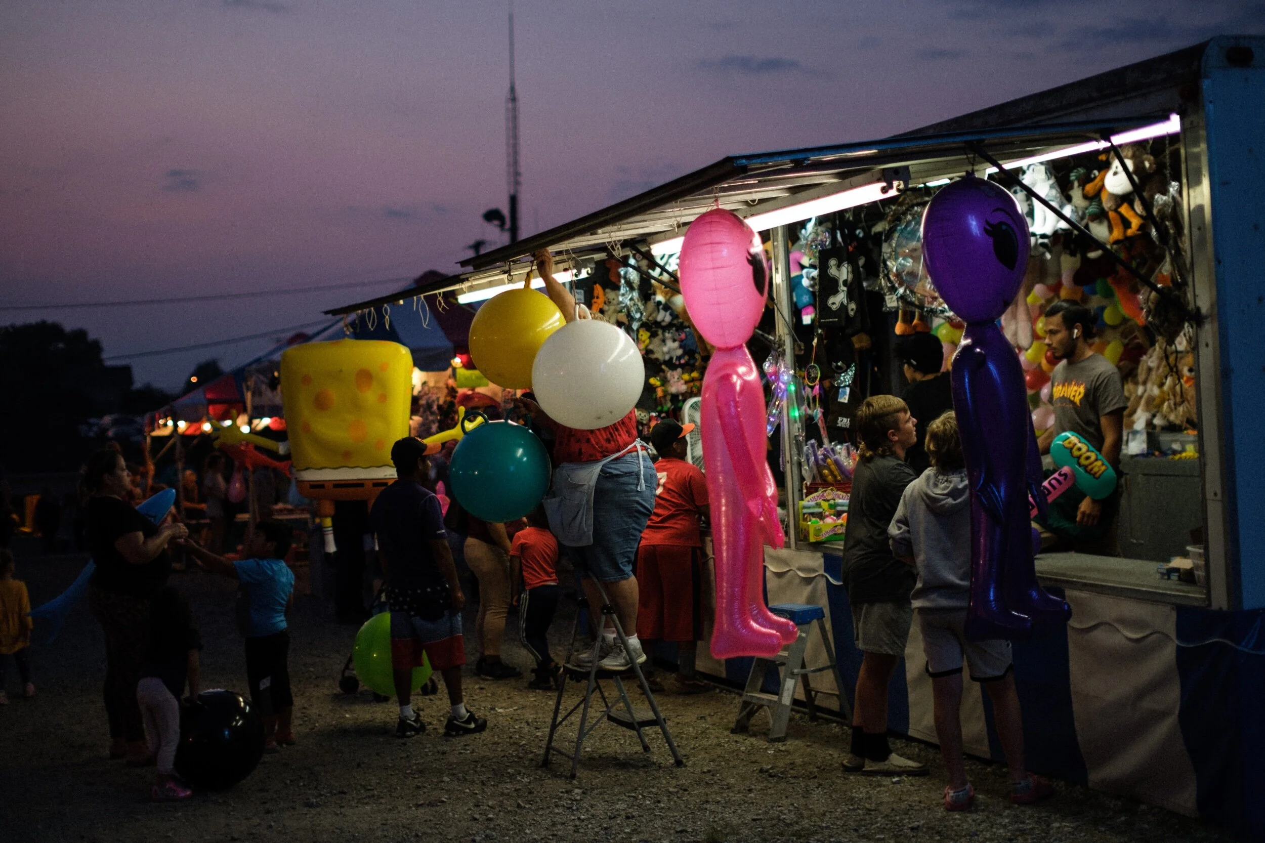 People at a nighttime carnival stand with prizes, including inflatable aliens, balloons, and plush toys.