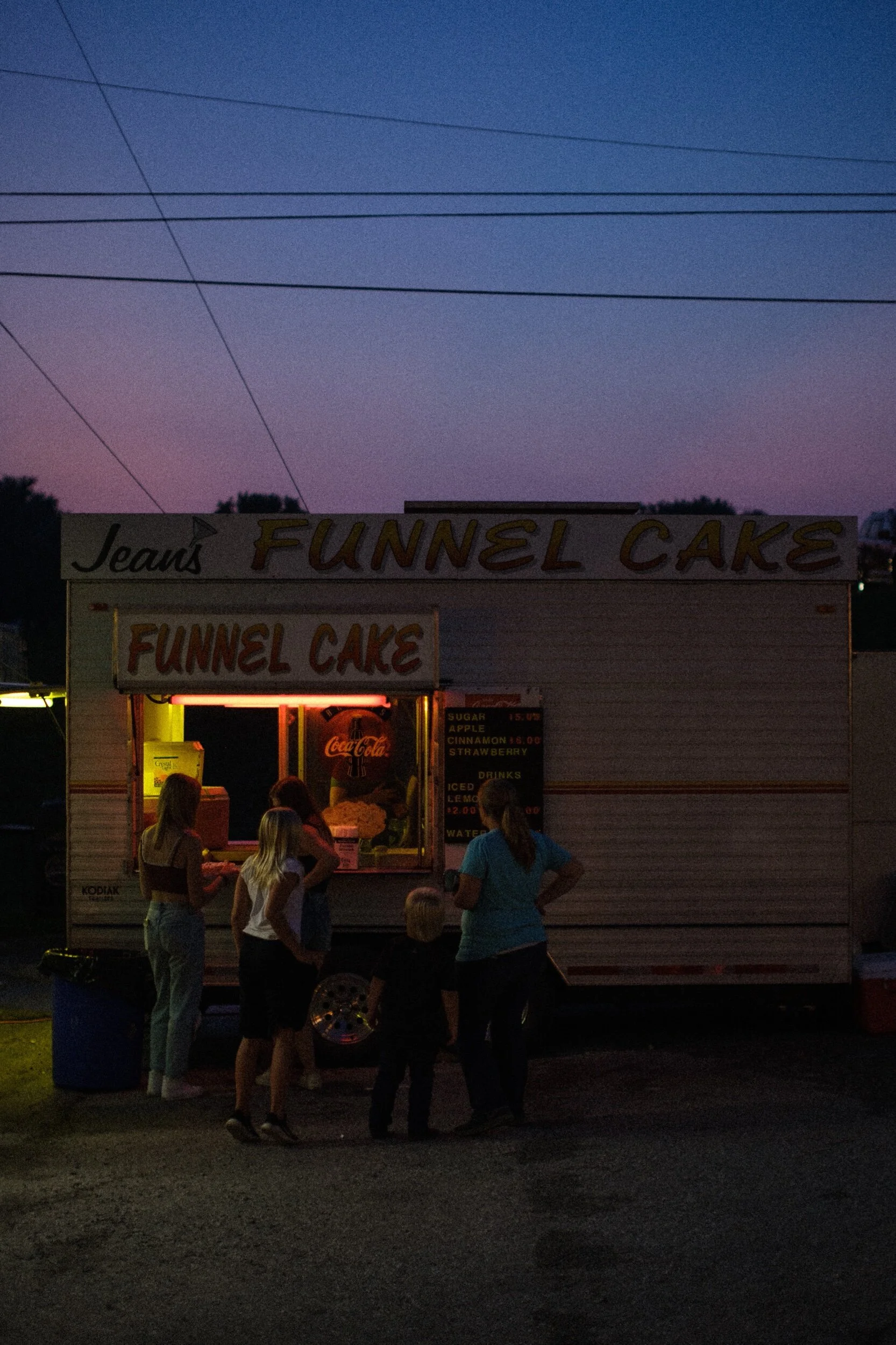 A group of people standing in front of a funnel cake food truck at dusk, with a sign that reads 'Jean's Funnel Cake.'