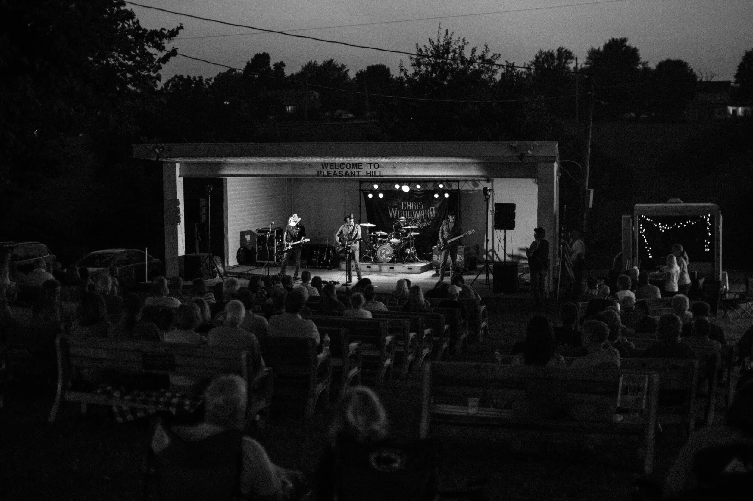 Outdoor concert at nighttime with audience seated on benches in front of a stage, featuring a band performing under a pavilion, trees silhouetted against the sky, and string lights in the background.