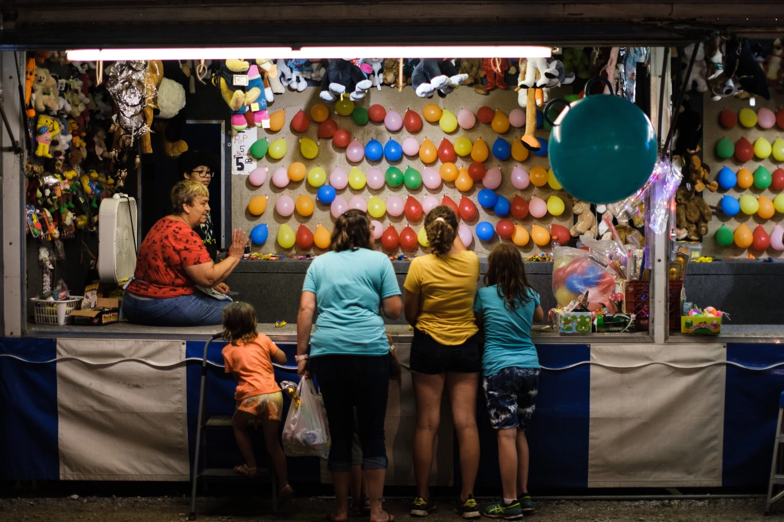 Children and adults playing a balloon dart game at a carnival booth with colorful stuffed animals as prizes.