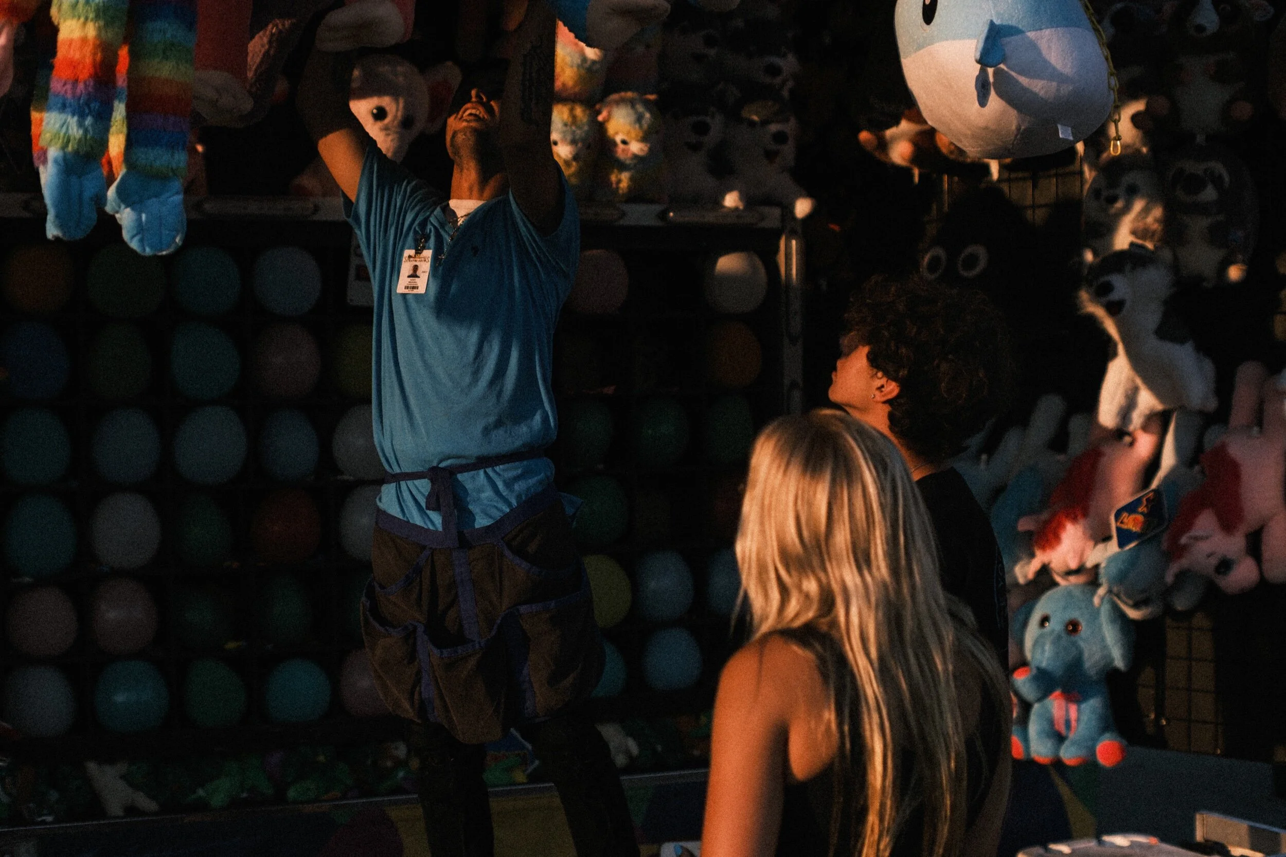Carnival game stand with a worker handing a stuffed toy to two visitors, surrounded by plush toys and a wall of colorful balloons.