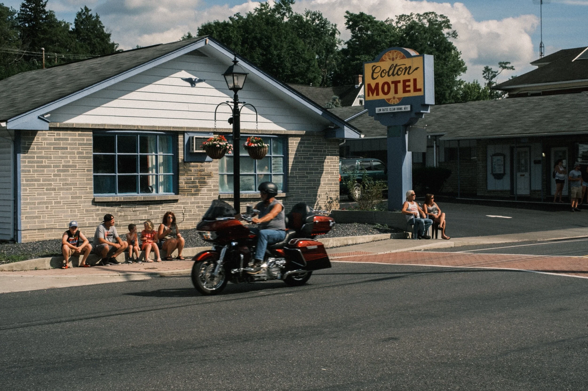 Motorcycle passing in front of a motel with people sitting outside, including a group on the curb and two people near the motel sign.