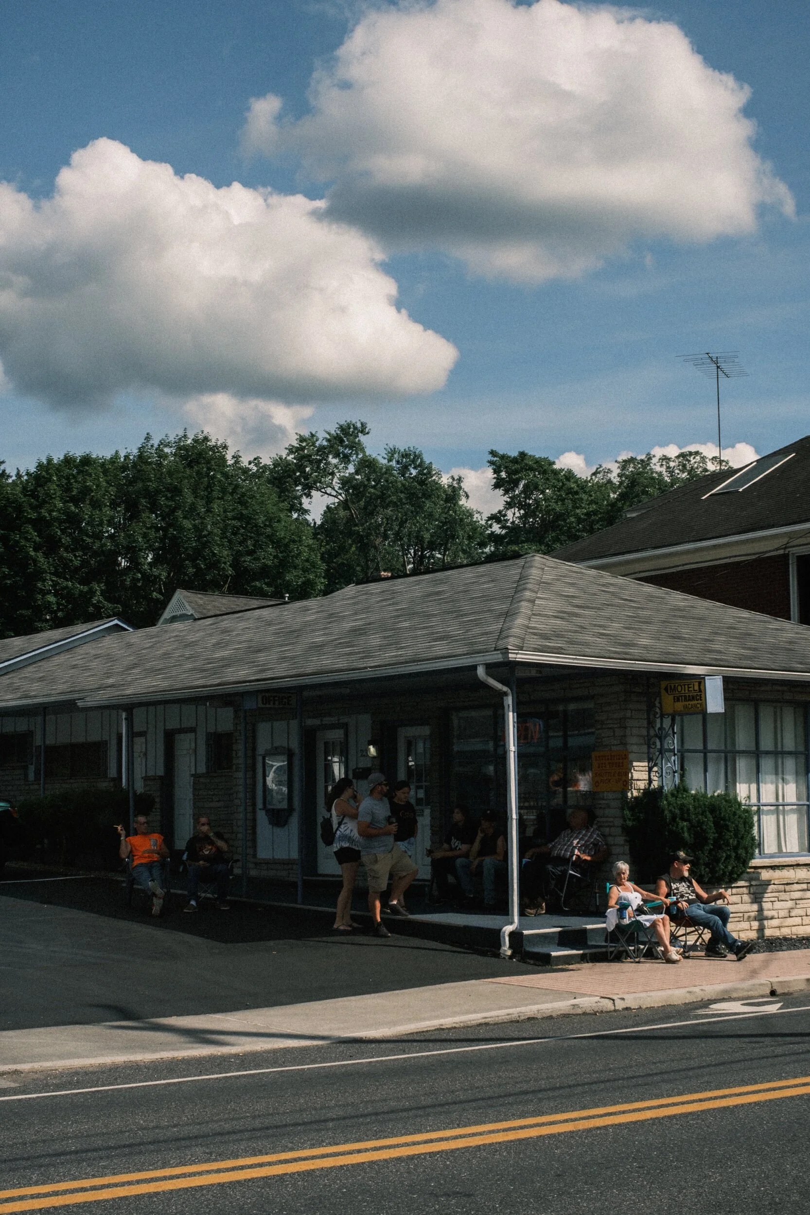 People sitting and standing outside a small building with a sign labeled "Motel Office," under a blue sky with clouds.