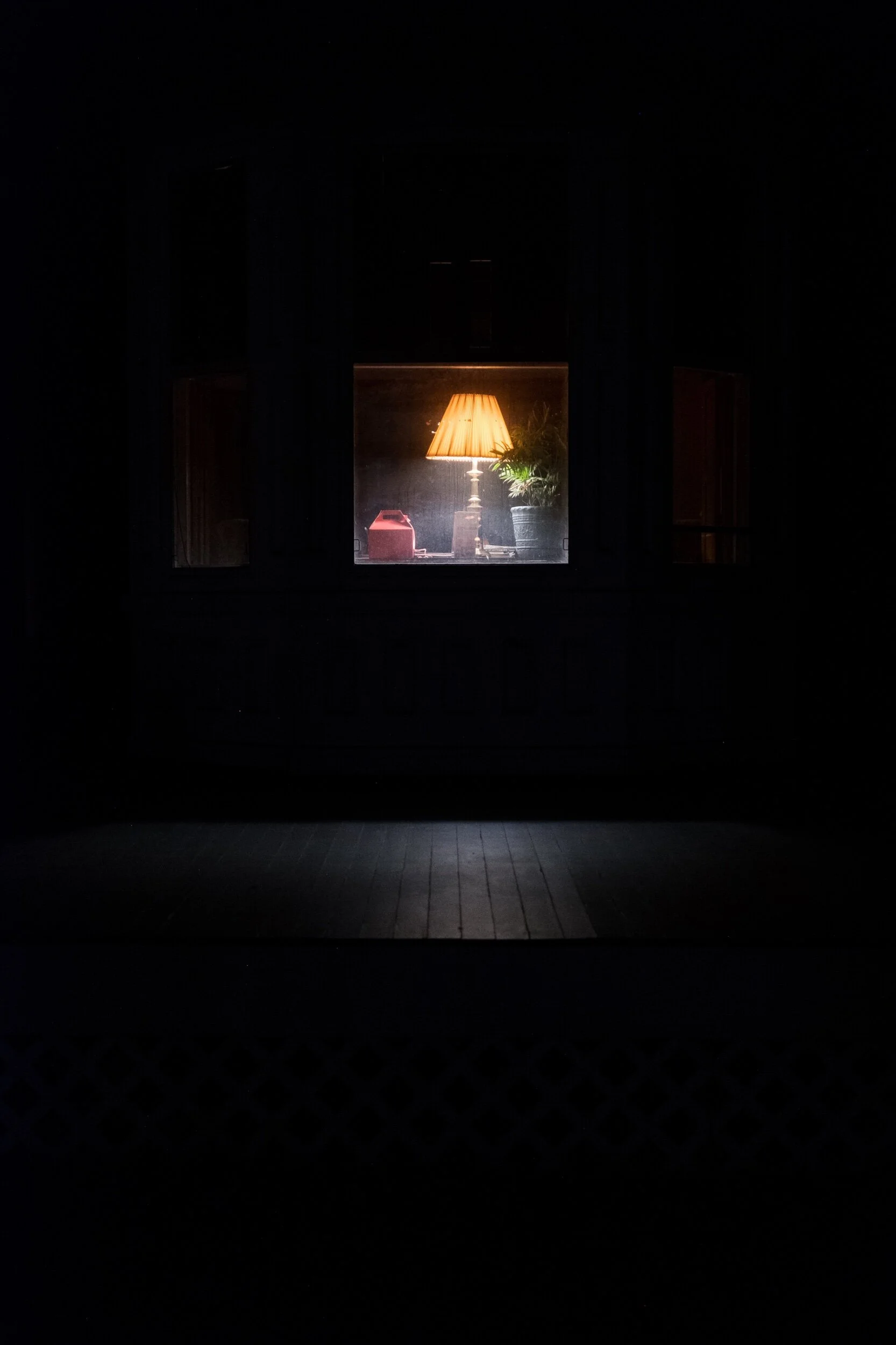 Dimly lit room with illuminated table lamp, red object, and plant visible through a window at night.