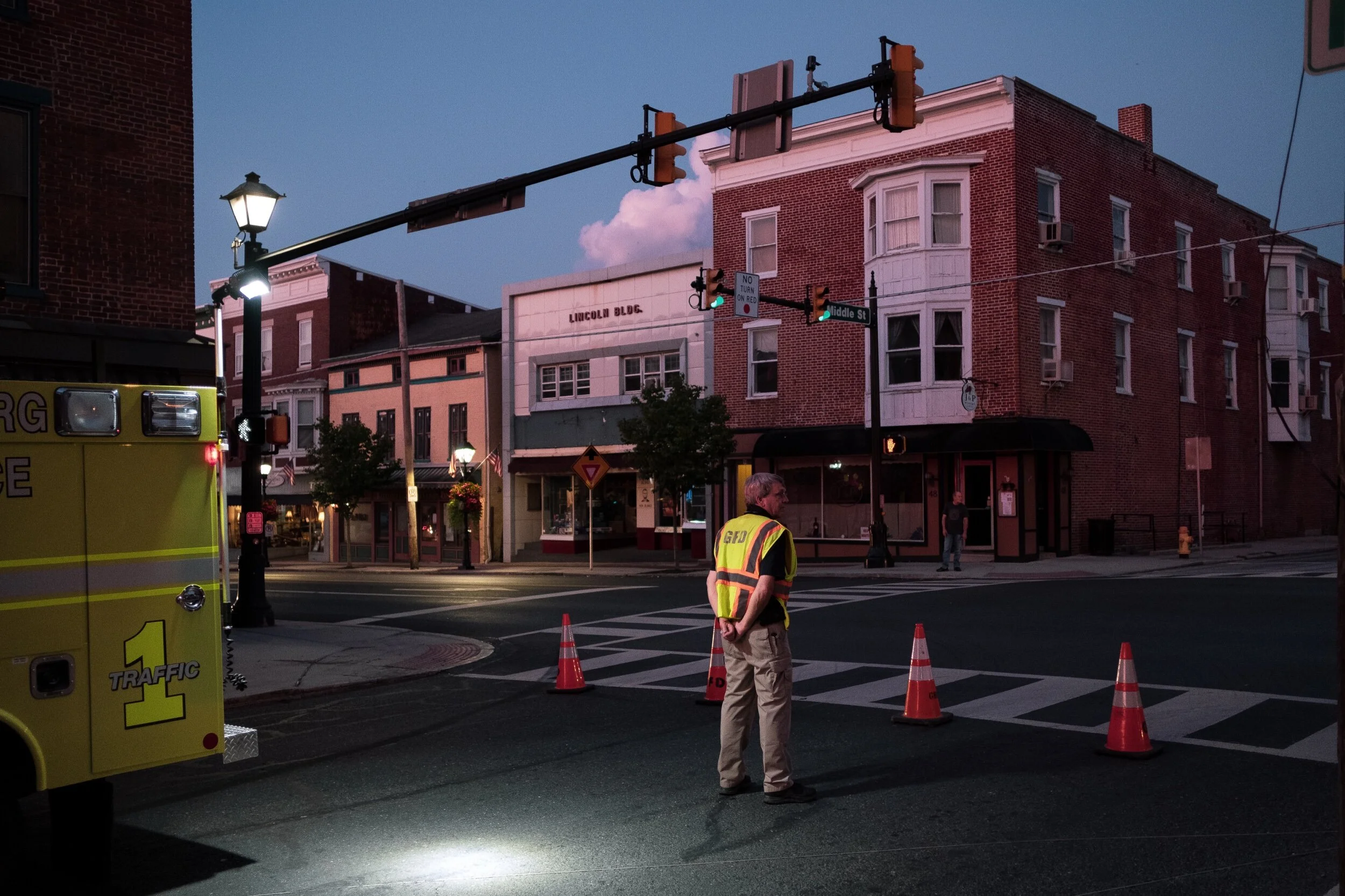 A nighttime street intersection with traffic cones and a man wearing a reflective vest, near a yellow emergency vehicle and lamp post, with brick buildings and shops in the background.