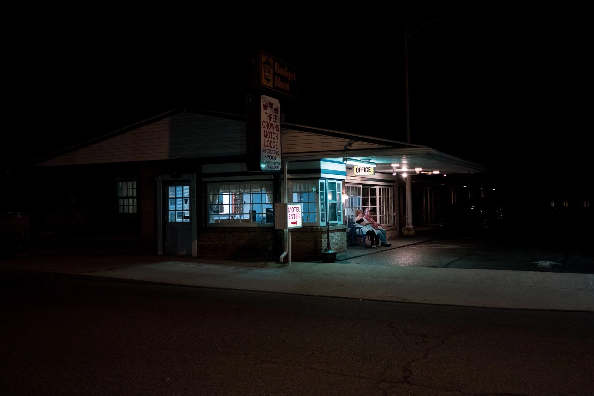 Exterior of a dimly lit motel office at night, with a glowing sign displaying 'Office'. A person sits on a chair near the entrance.