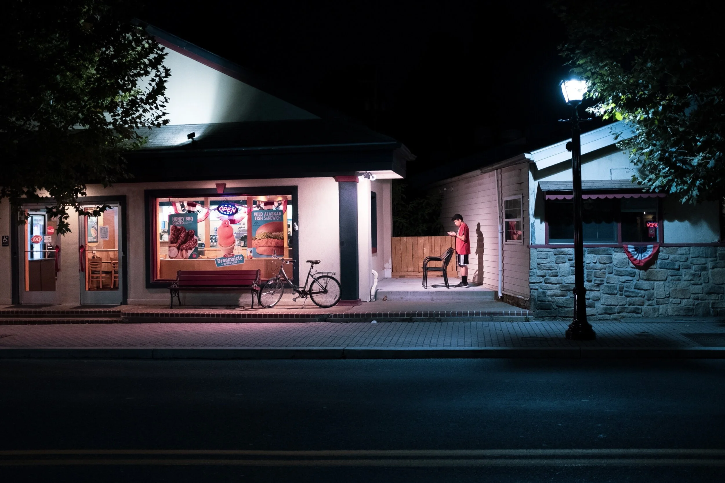 Street view of an ice cream shop at night with a neon open sign, a bicycle parked outside, and a person in a red shirt standing on a patio using a phone.