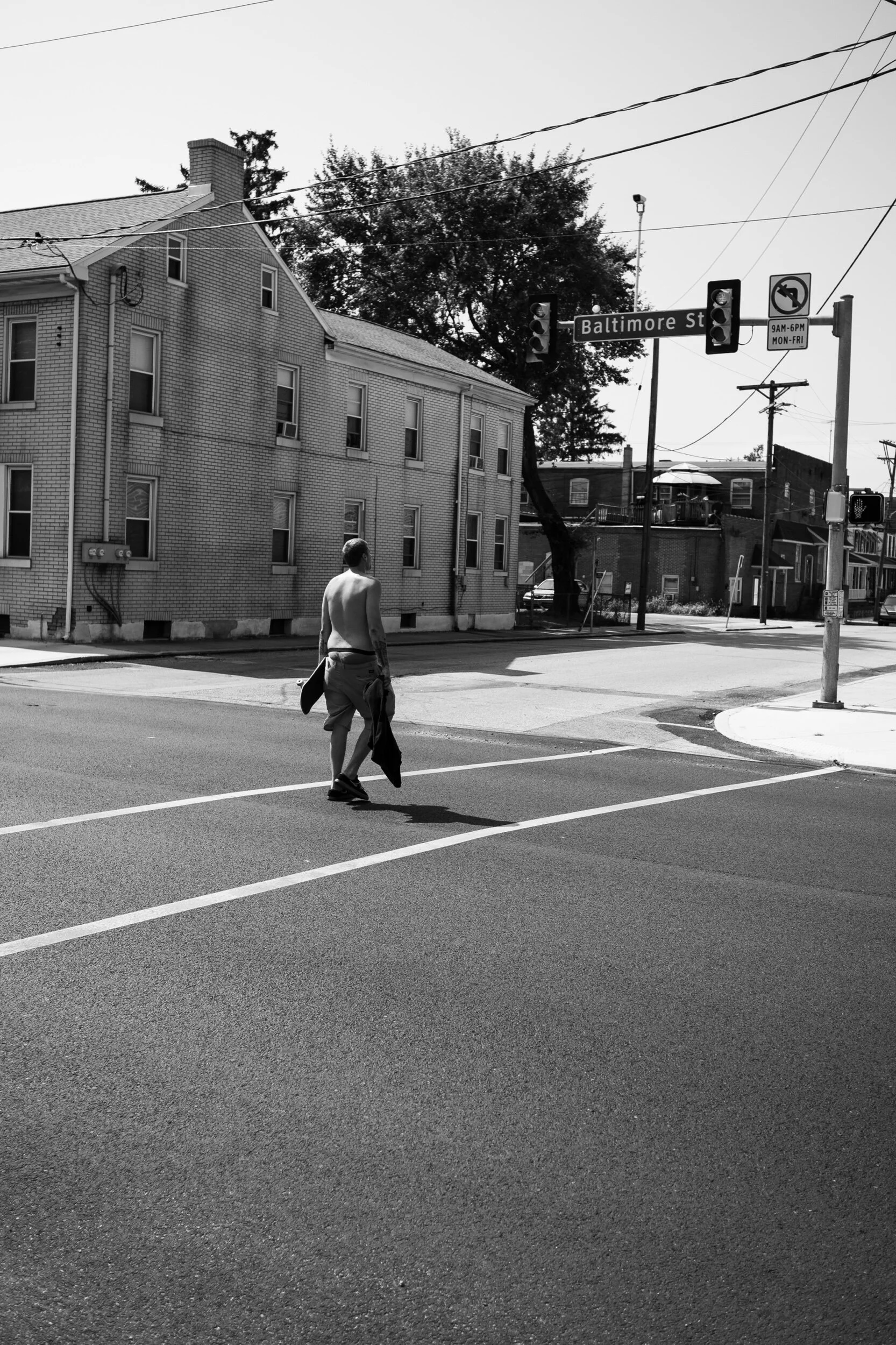 Black and white photo of a shirtless man walking across a street intersection near Baltimore St, with residential buildings and traffic lights in the background.