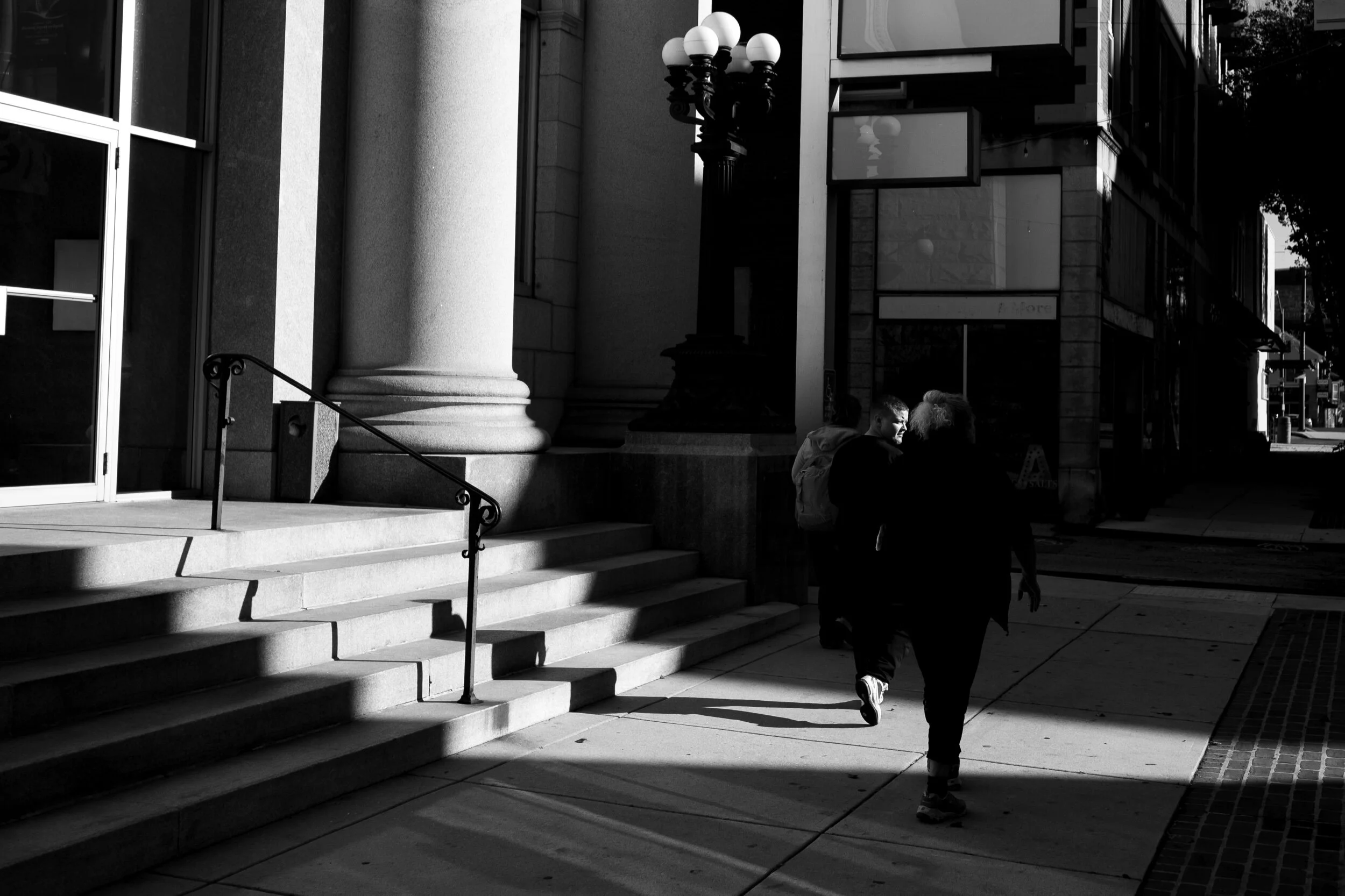 Black and white photo of people walking past large building columns and stairs, with street lamps and shadows.