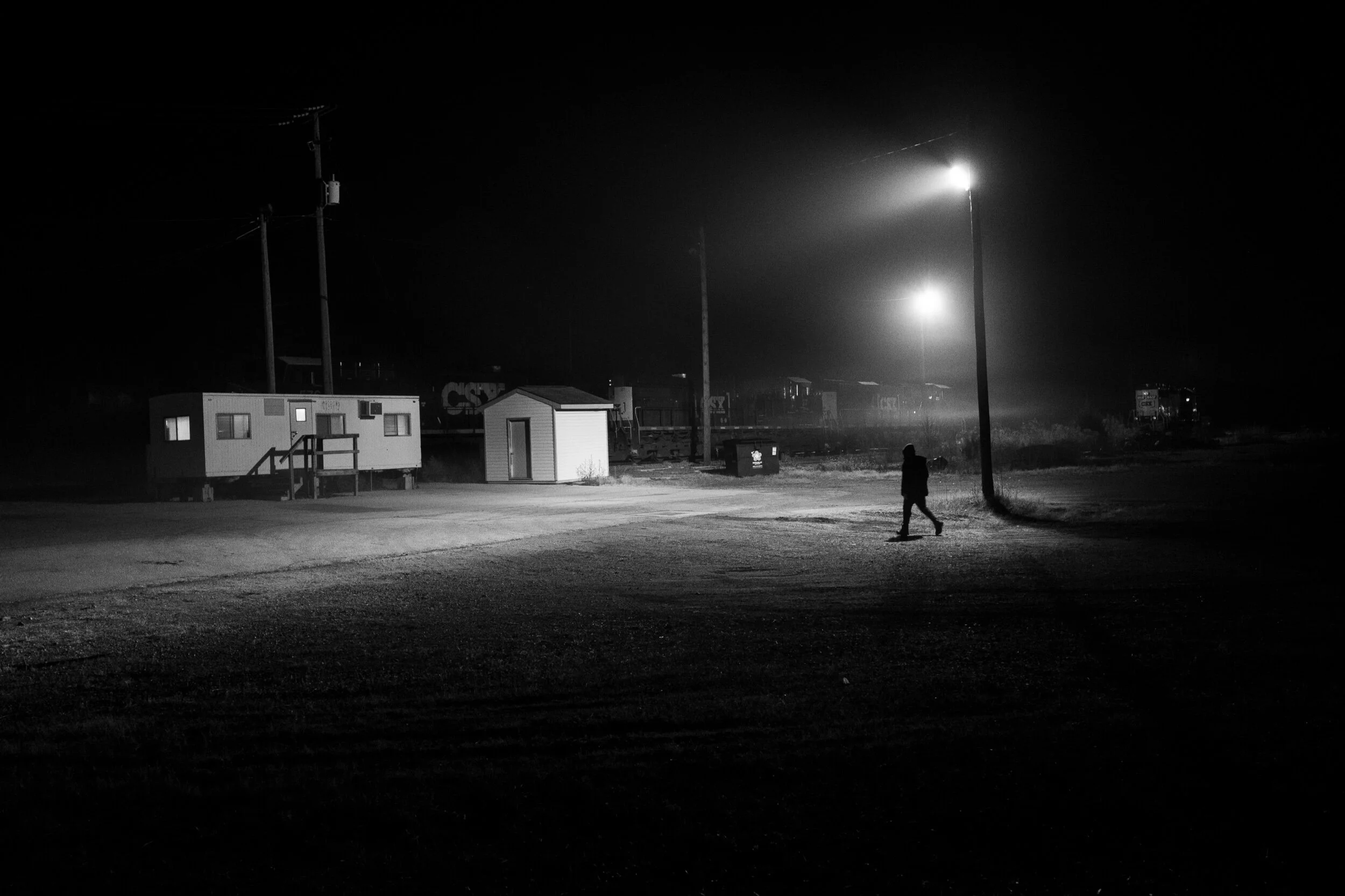 Black and white photo of a person walking in a dark industrial area with a trailer and a shed, illuminated by streetlights.
