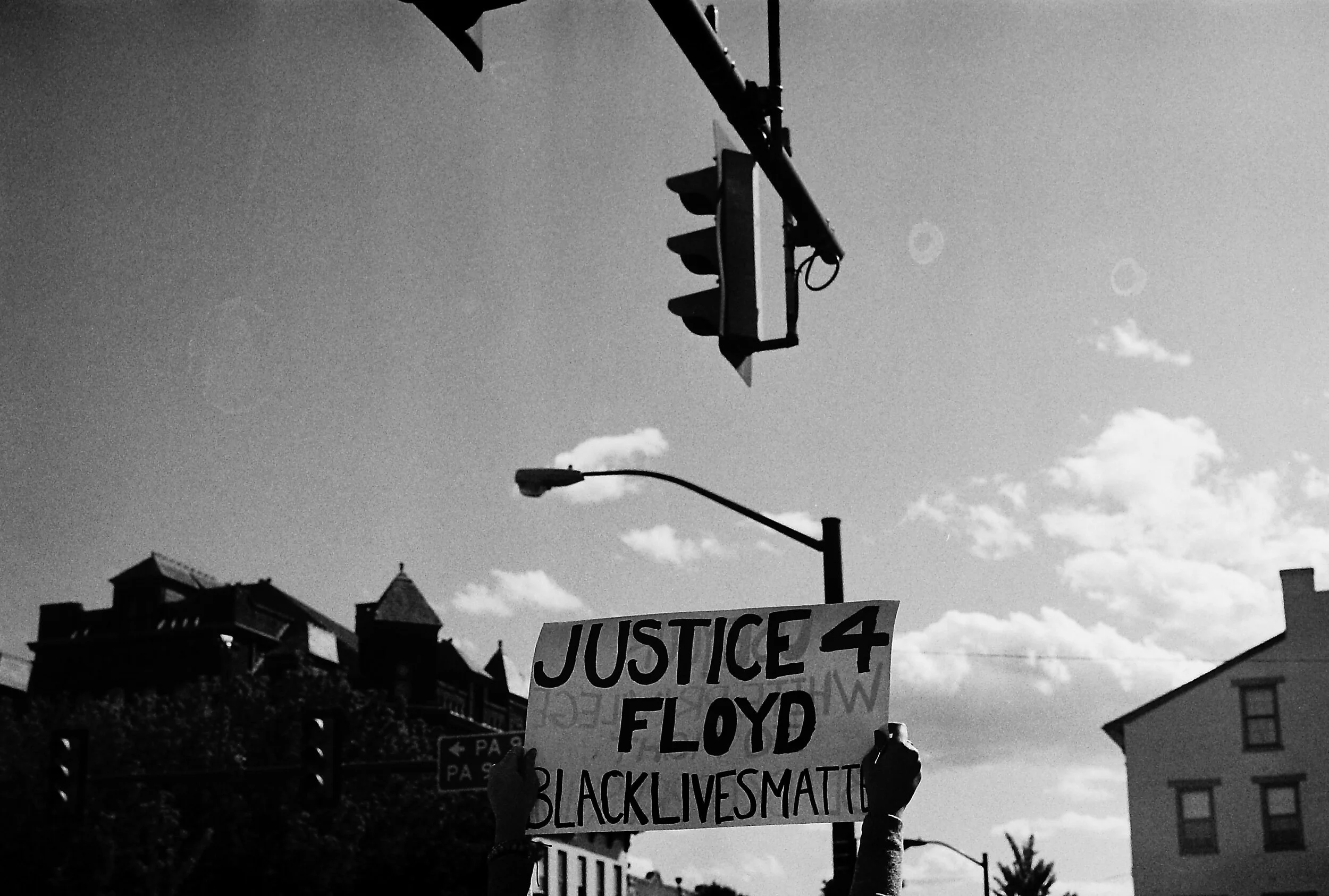 Black and white photo of a protest with a person holding a "Justice 4 Floyd Black Lives Matter" sign under traffic lights.